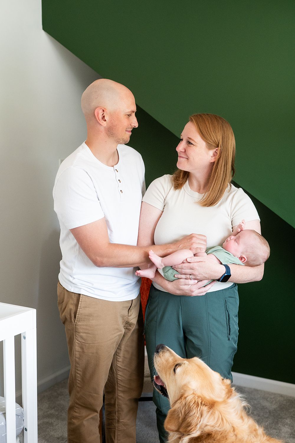Couple stands together with baby in green room while golden retriever looks on attentively.