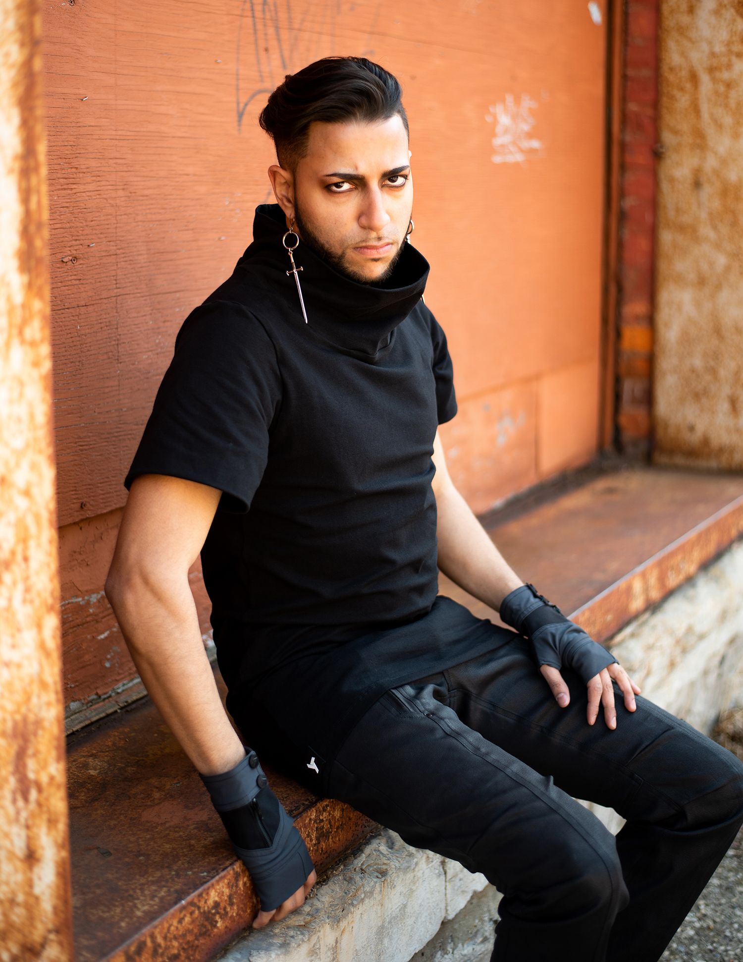 Person in an all-black outfit with earrings sits against an orange wall in an edgy fashion pose.