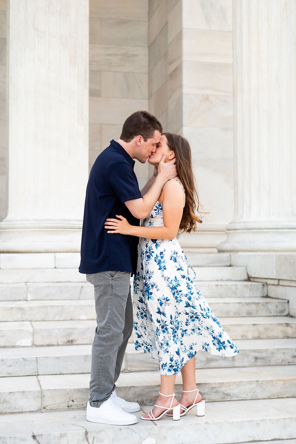 Romantic couple sharing a kiss on marble steps, with her wearing a blue floral dress and him in a navy shirt.