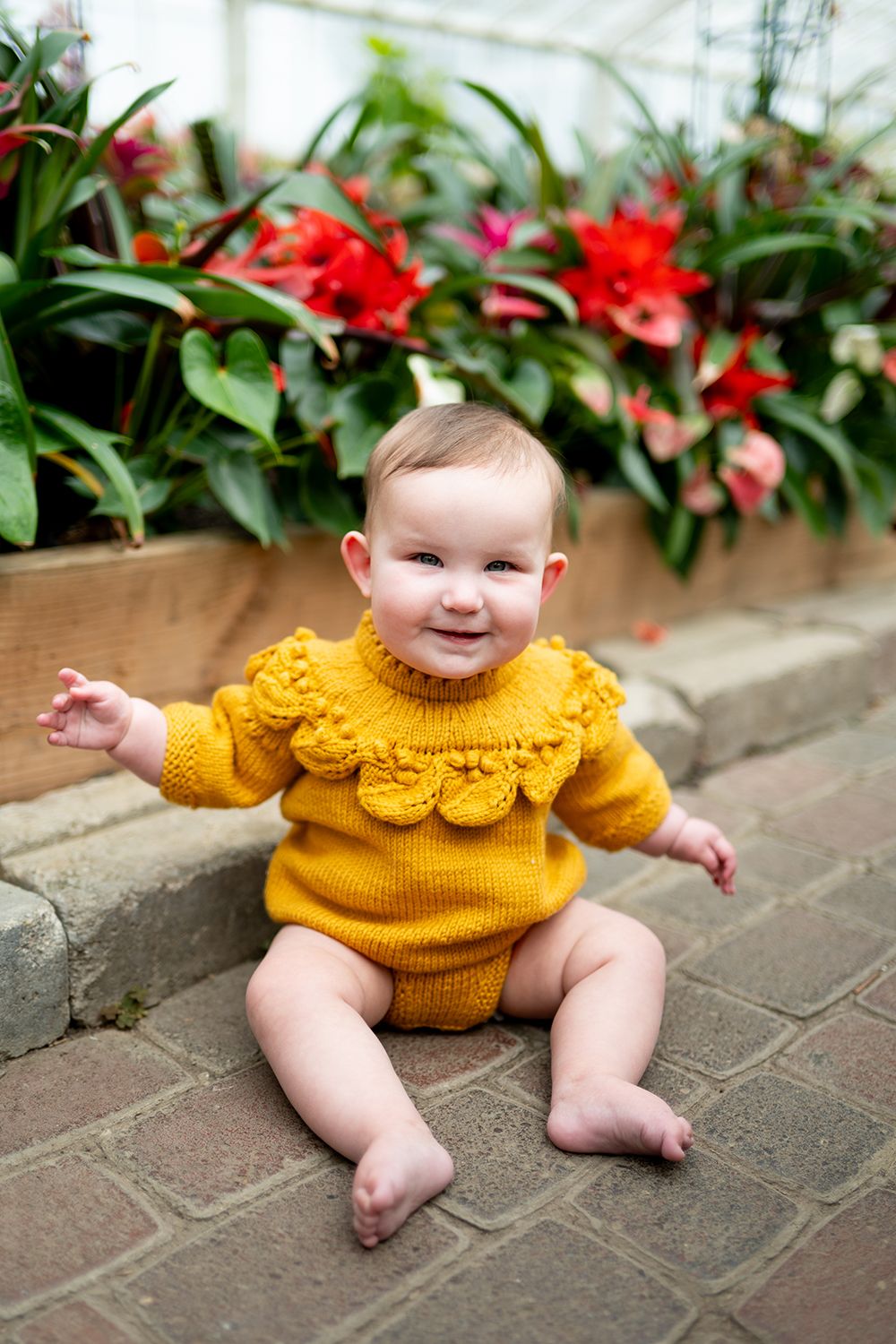Baby in bright yellow sweater smiles while sitting on brick pavers beside flowering plants.