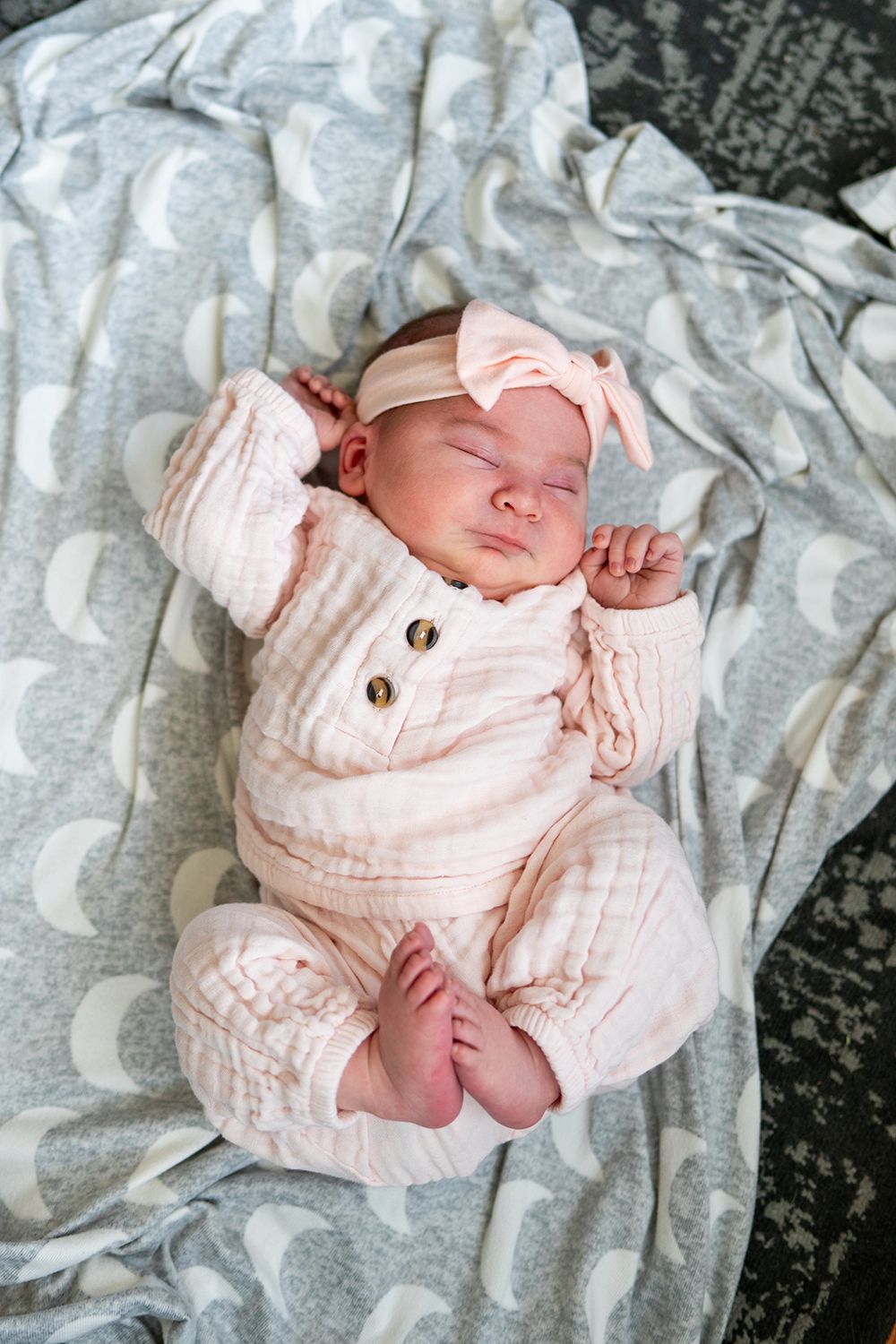 Sleeping newborn in pink buttoned outfit stretches while lying on gray patterned blanket.