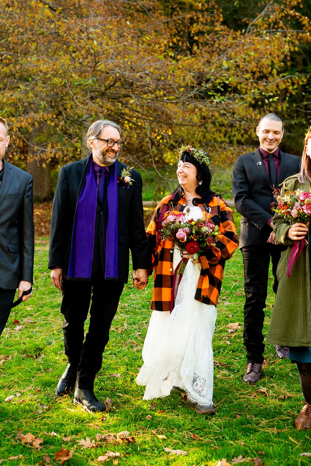 Wedding party wearing autumn attire stands on grass with fall foliage in background during outdoor celebration.