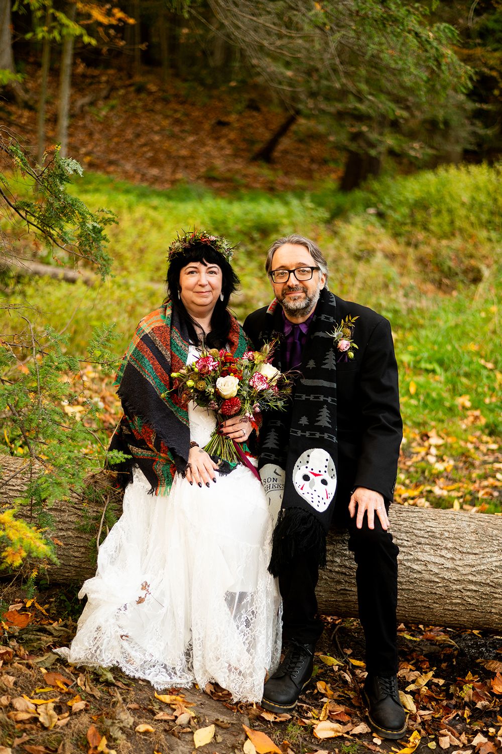 Two people sit together on a fallen log in the woods wearing Halloween-themed wedding attire.