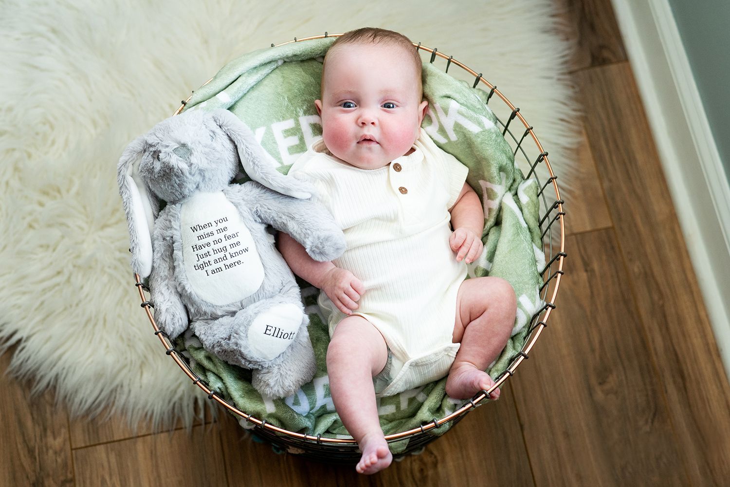 A small infant in white onesie lies in a round basket on top of a fluffy rug with a stuffed animal beside them.