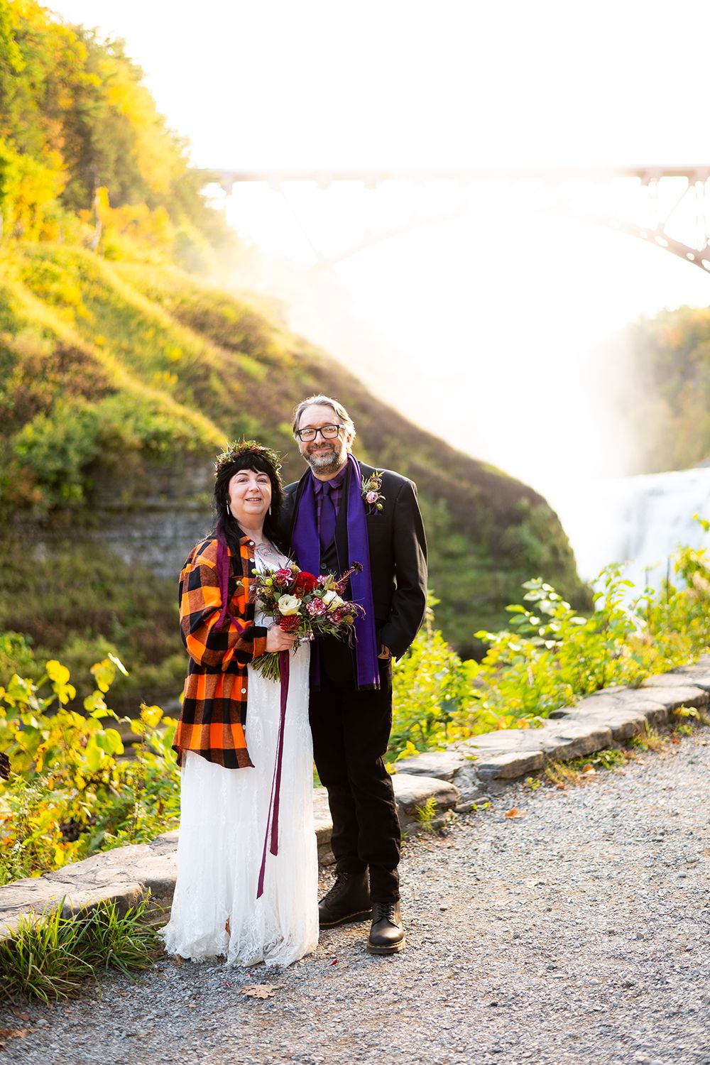 Two people stand on a scenic trail near waterfalls and autumn foliage at sunset, one wearing a plaid jacket and holding flowers.