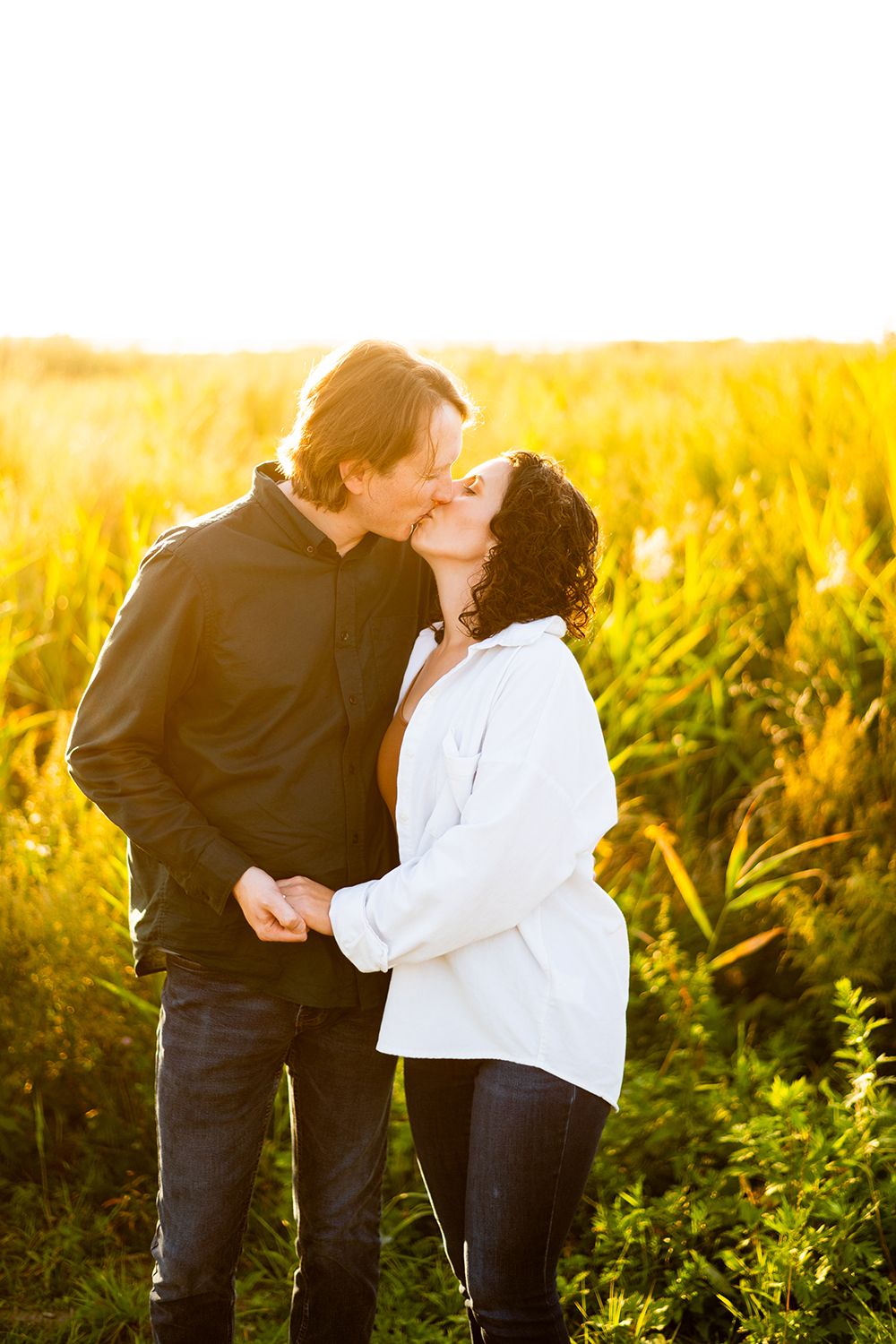 A couple shares a romantic kiss in a golden sunlit field during sunset, with warm glowing light around them.