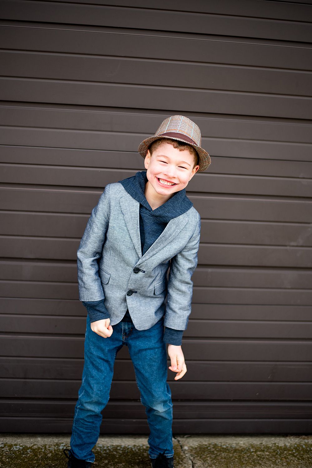 A child in a gray blazer, hat and jeans stands against a dark wall with a happy expression.