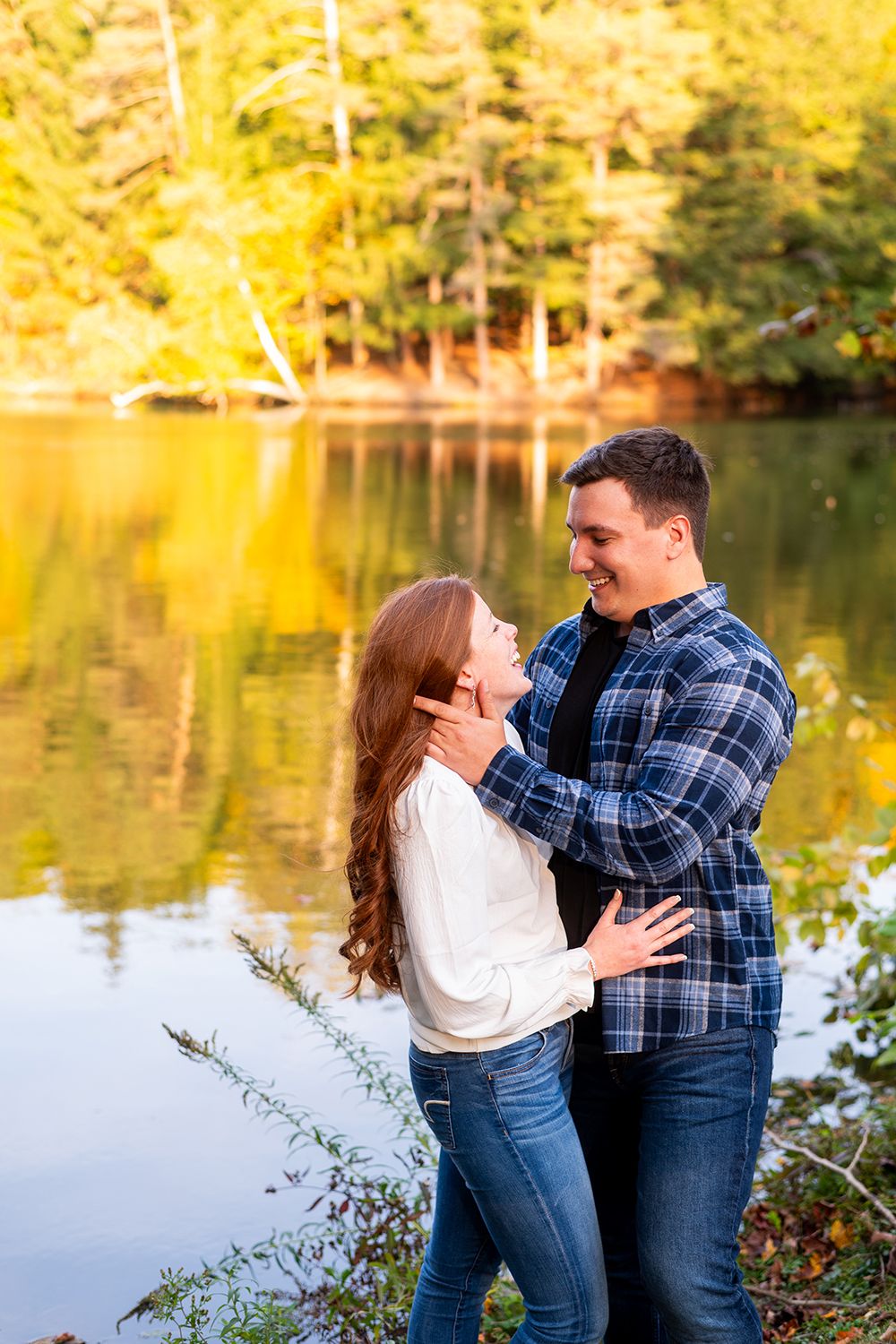 Couple in plaid shirt and white sweater sharing romantic moments near a lake reflecting golden autumn foliage.