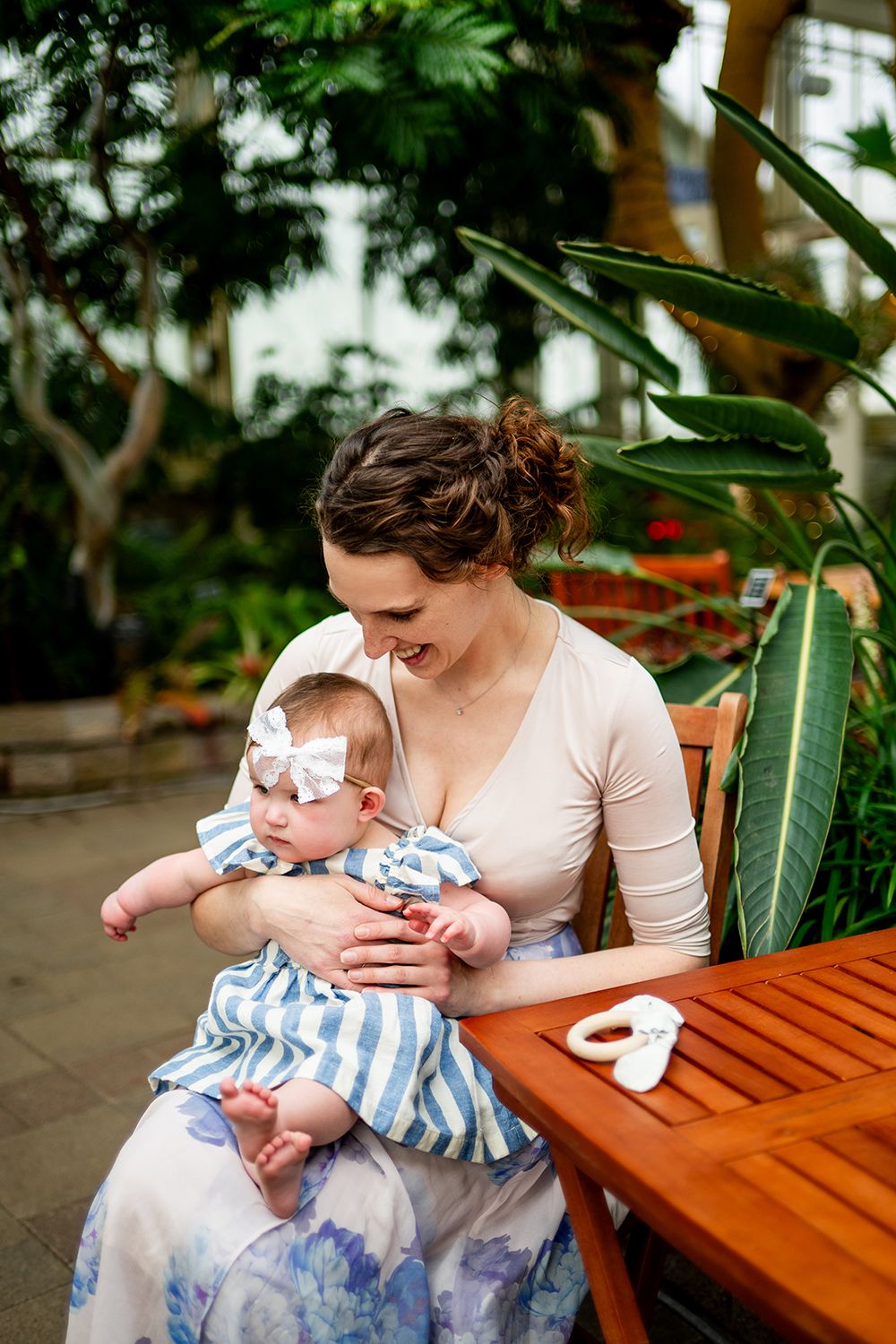 A tender moment between mother and baby sitting at a wooden table in a greenhouse setting amid lush plants.