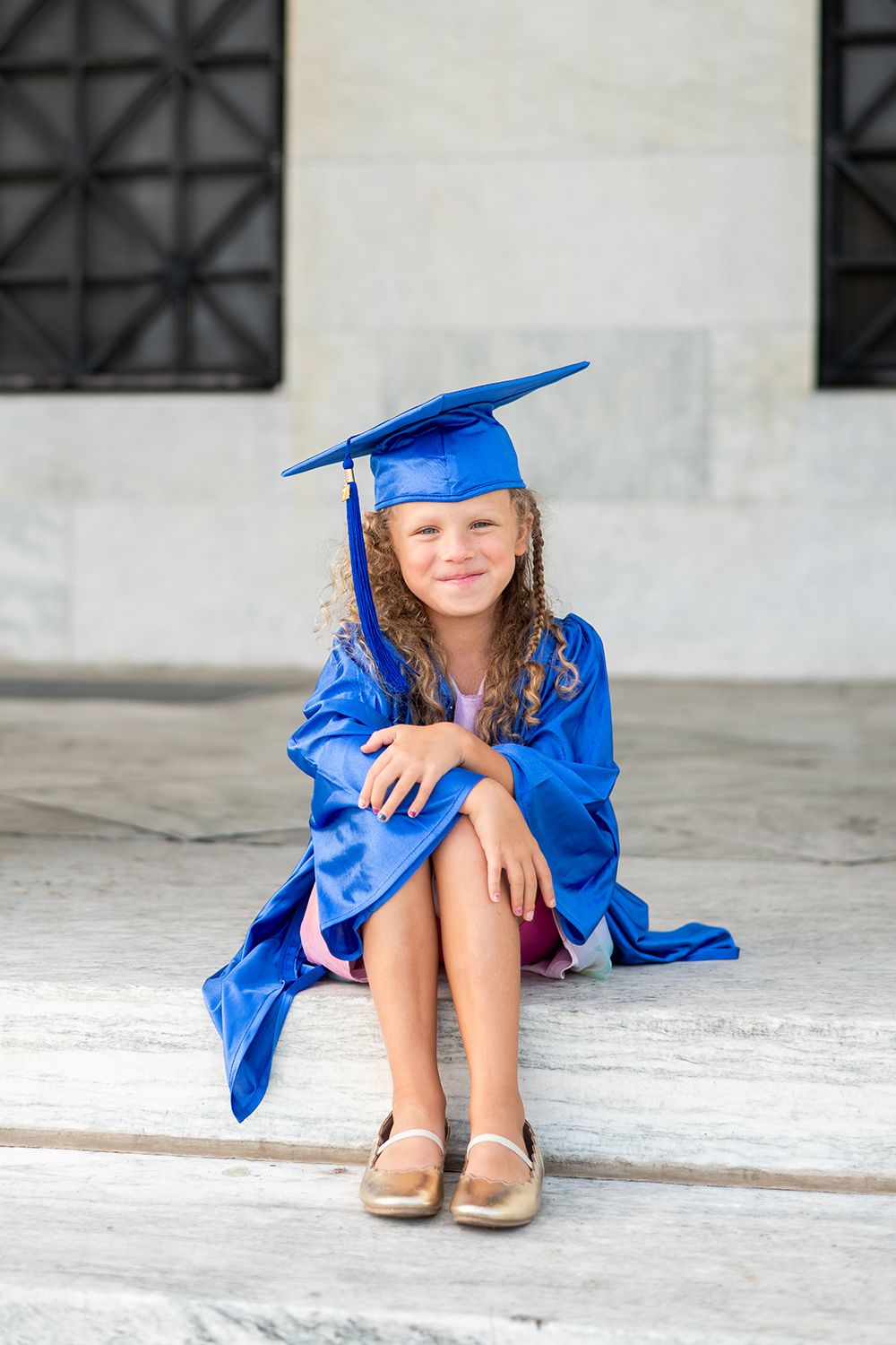 Young graduate in bright blue cap and gown sits on stone steps wearing beige shoes.