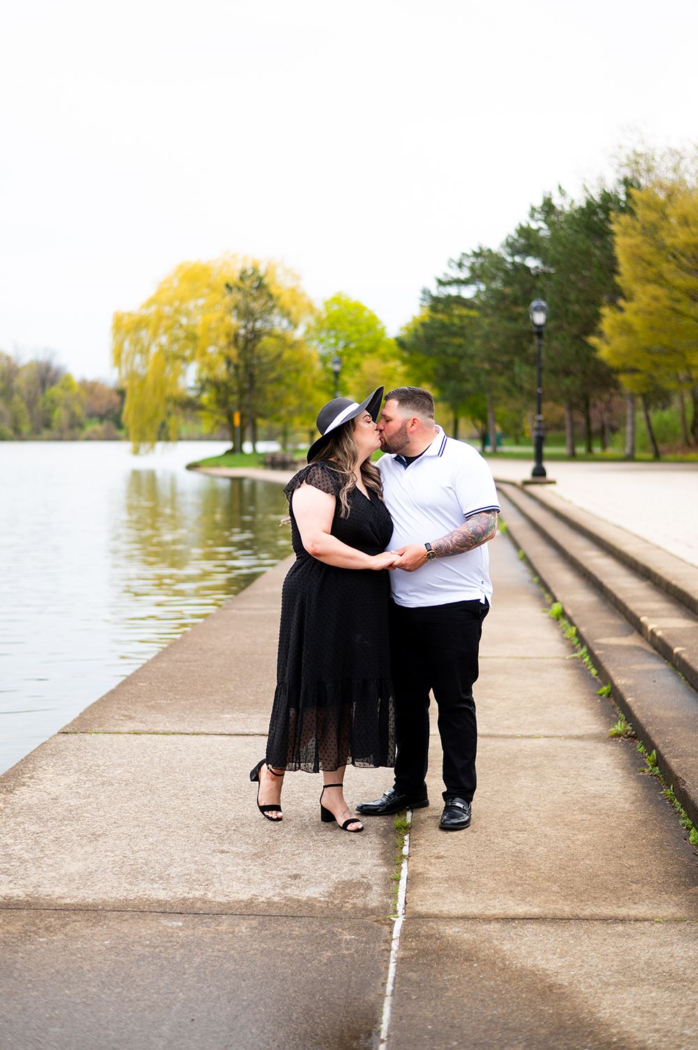 A romantic couple in formal attire sits together on concrete steps near a peaceful waterside promenade in autumn.