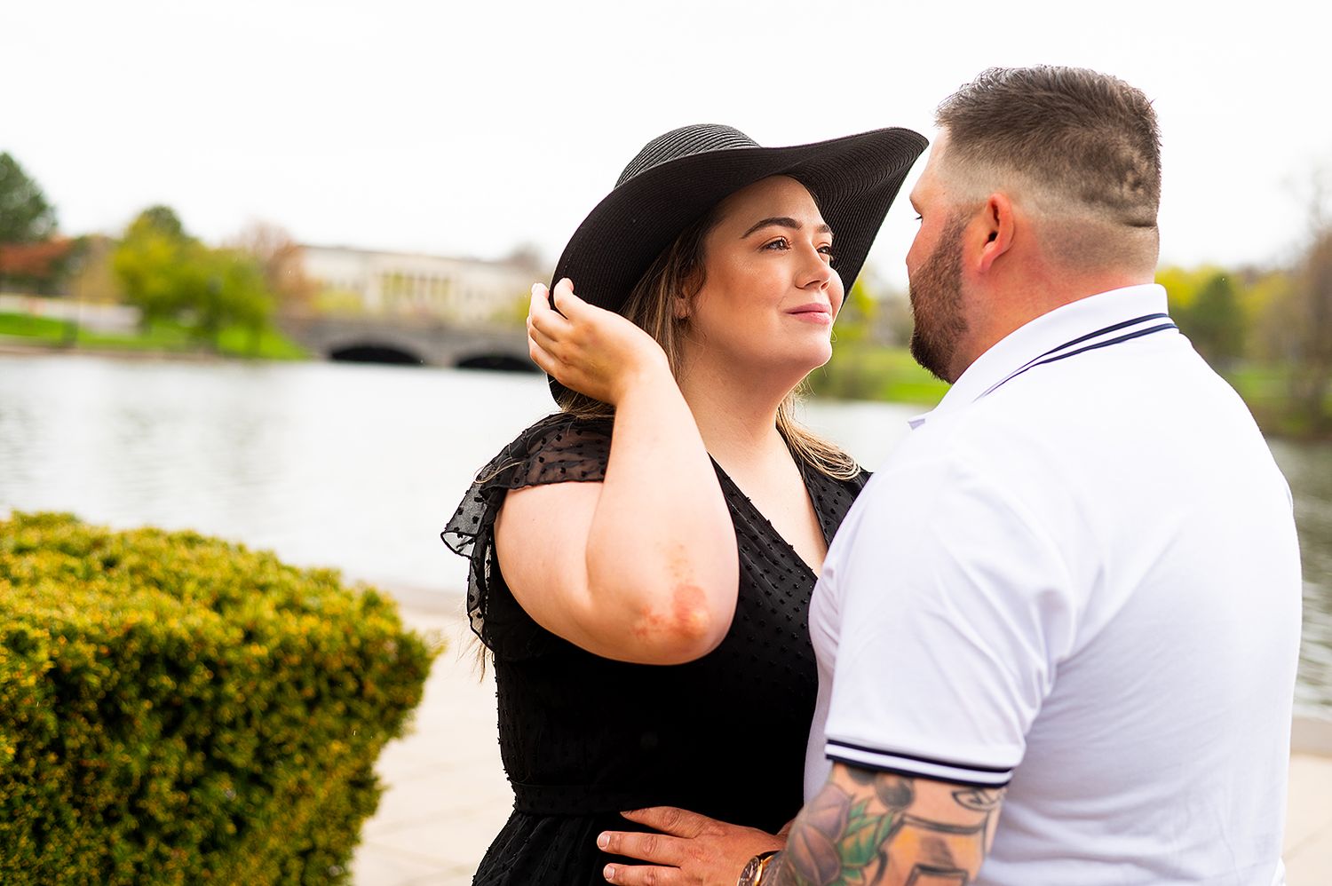 A couple shares an intimate moment outdoors by landscaped hedges, with her adjusting a wide-brimmed black hat.