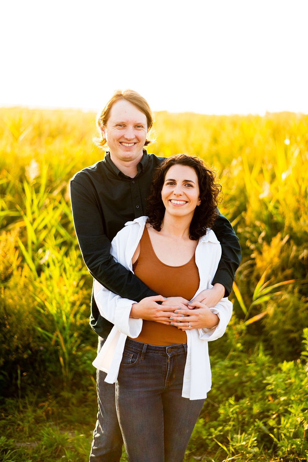 A couple embraces in a golden field during sunset, sharing a warm moment among tall grass and sunlight.