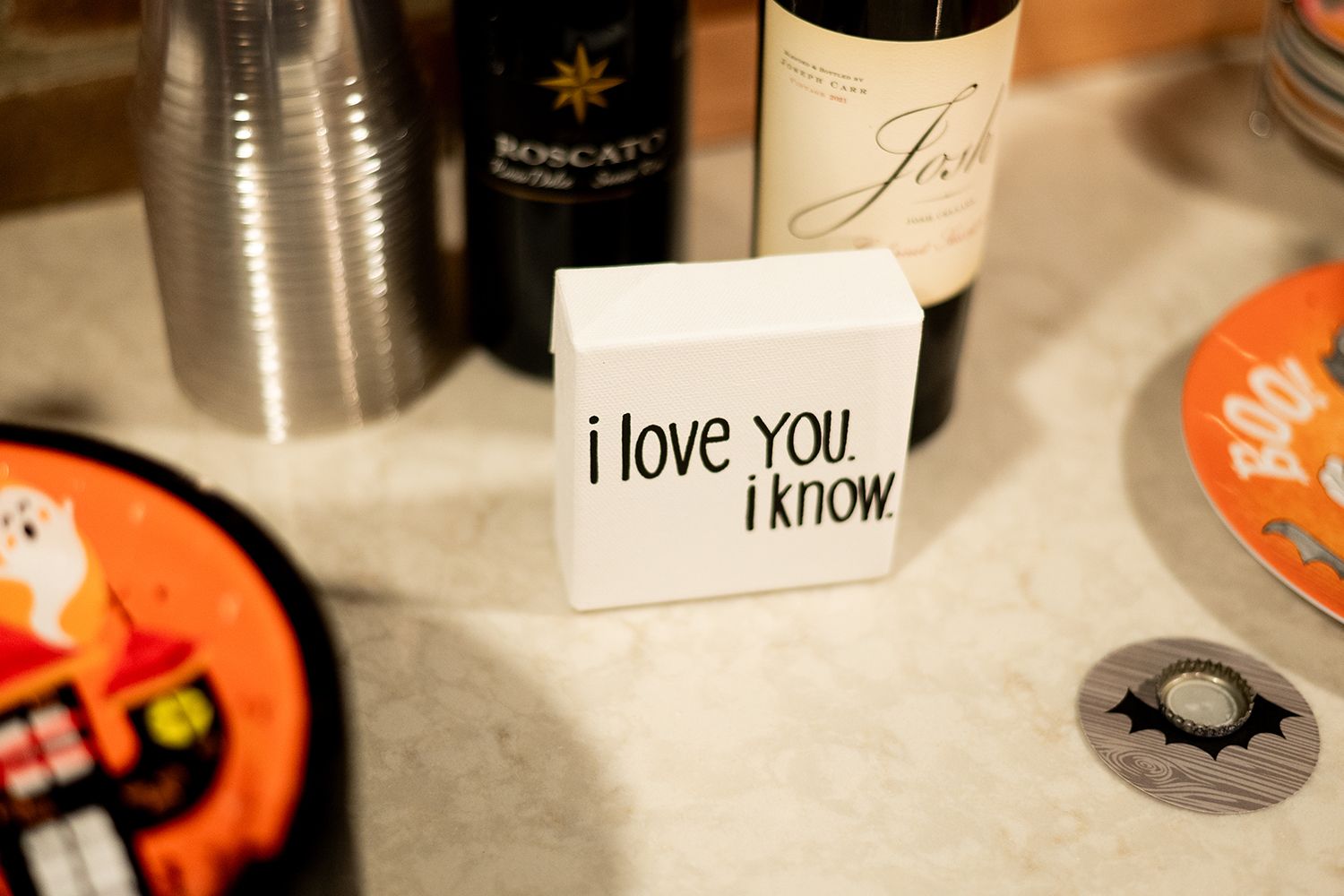 A handwritten note reading 'i love you i know' sits on a table next to wine bottles and Halloween decorations.