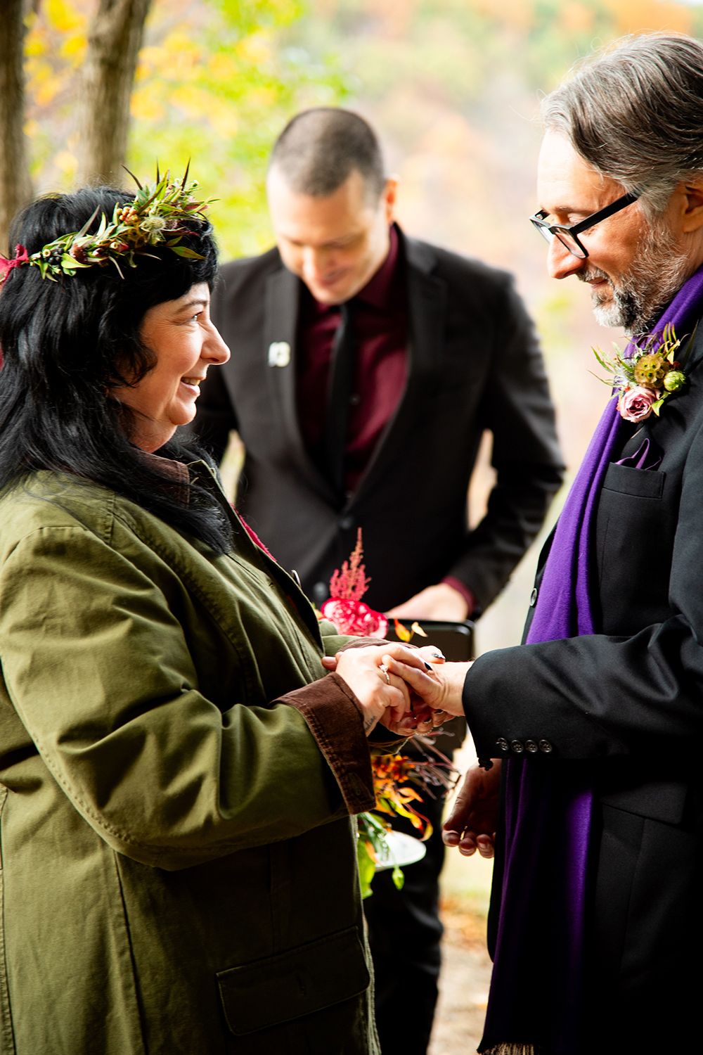 Couple exchanges rings during an outdoor autumn wedding ceremony with an officiant in a purple stole present.