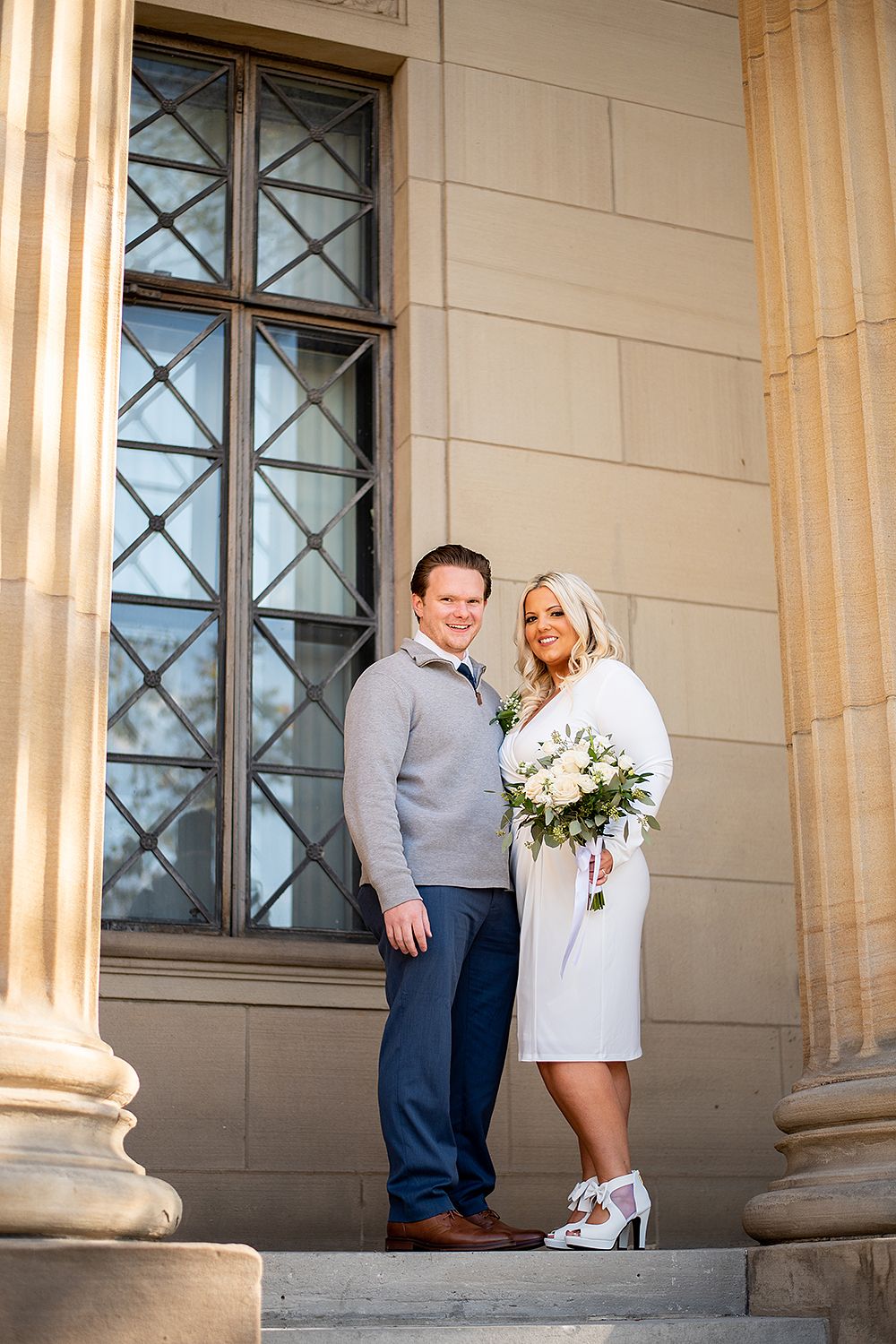 Couple poses together on stone steps between classical columns with an elegant white floral bouquet.