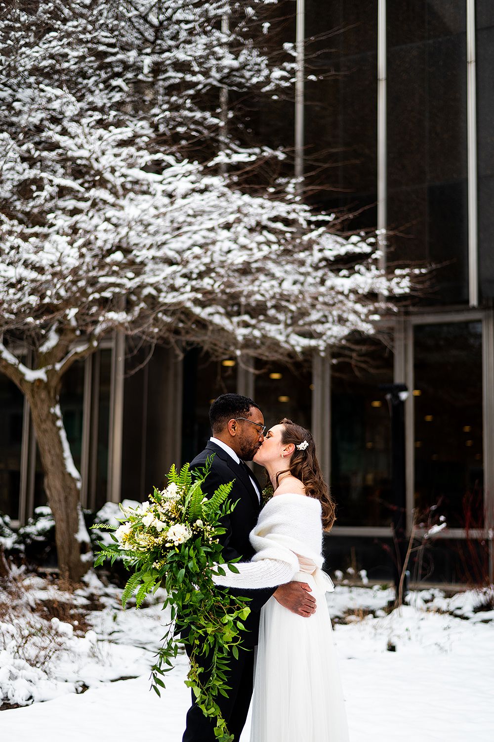 A winter wedding couple embraces under a snow-covered tree outside a modern building.