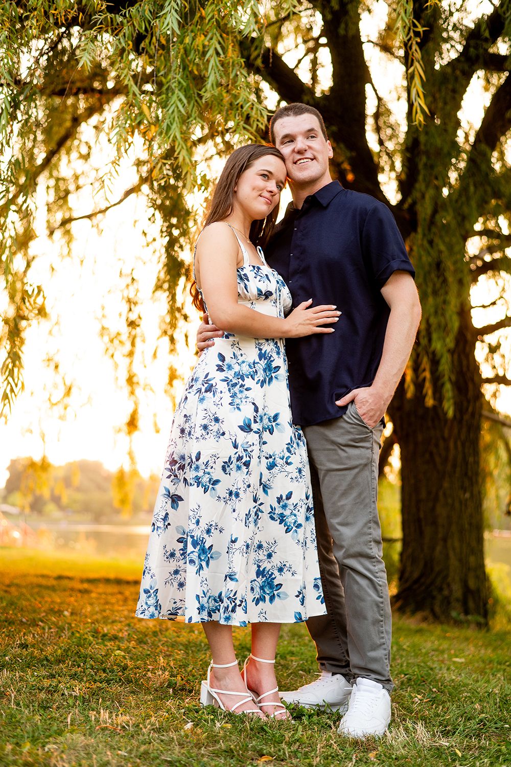 A couple embraces while standing beneath willow trees at sunset in a park setting.