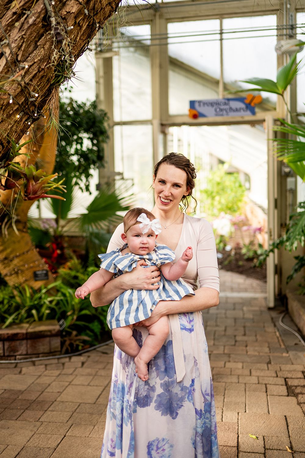 A woman in a floral dress holds a baby wearing blue and white stripes inside a greenhouse with brick flooring.