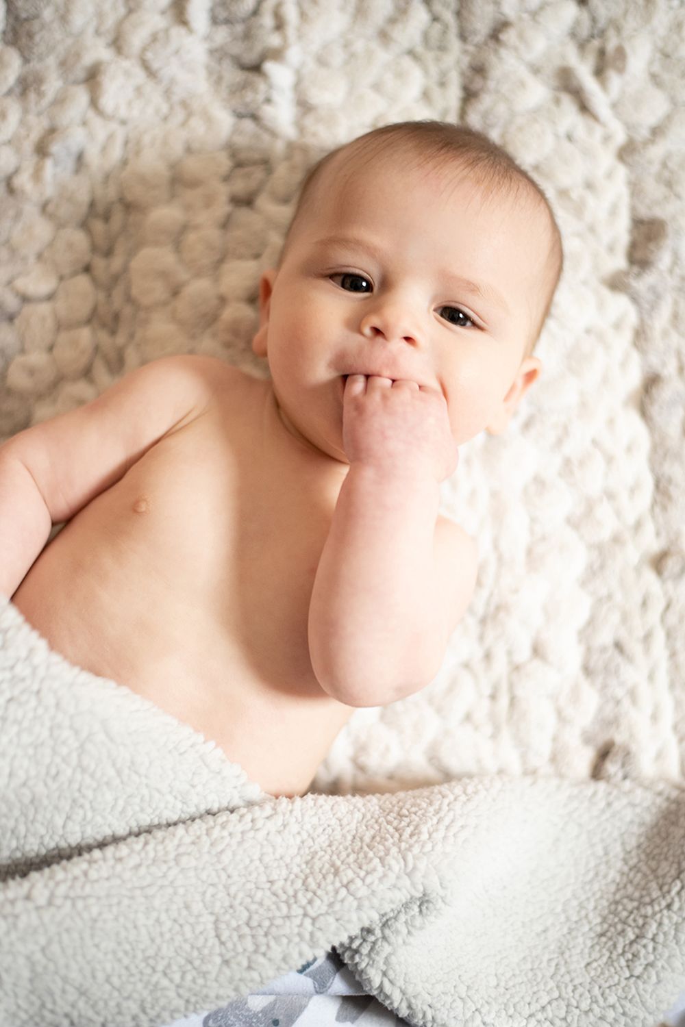 An infant lies on soft textured bedding with a playful expression while sucking on their fingers.