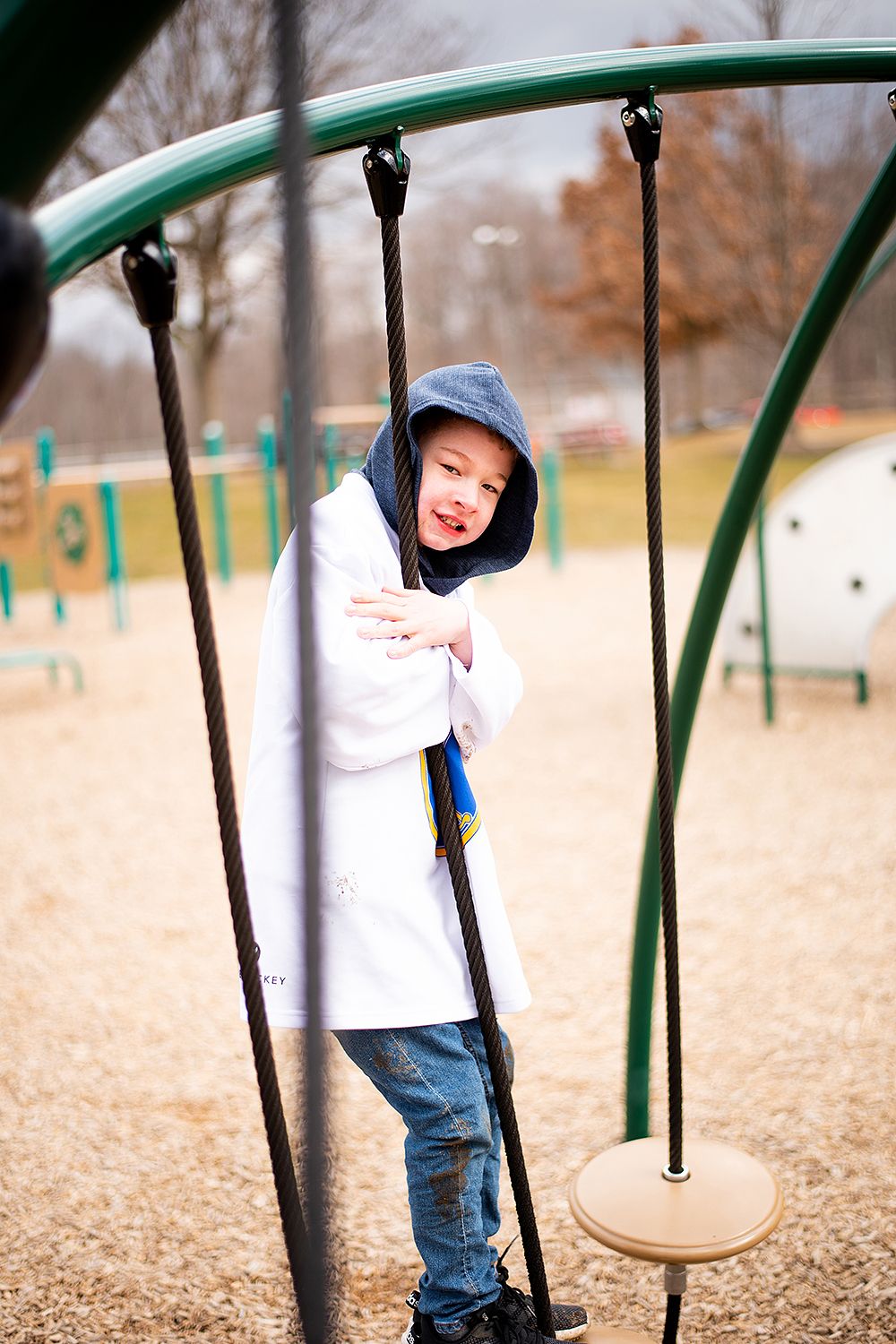 A child in a white hoodie and blue cap plays on playground equipment with metal poles and a disc platform.