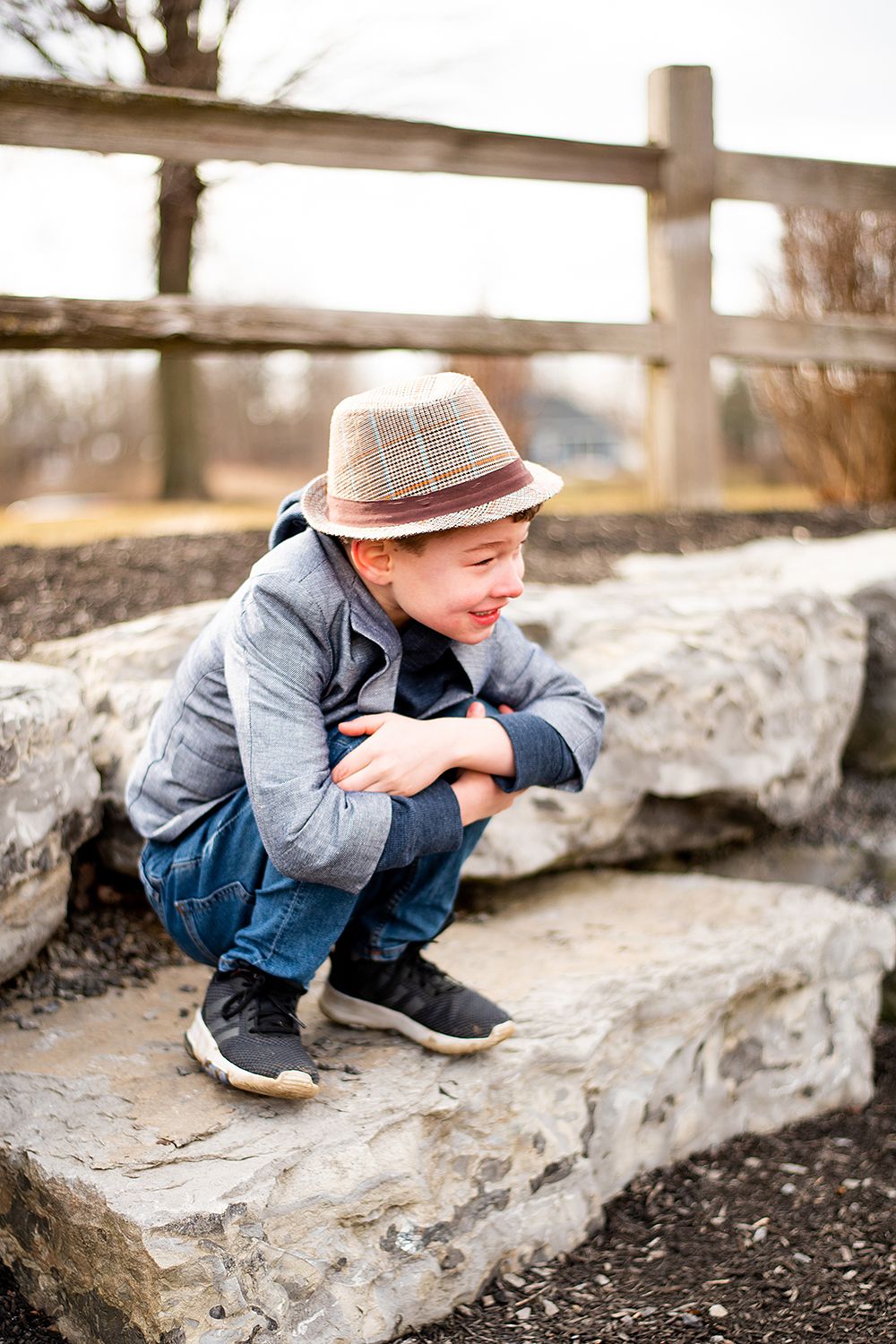 Child in fedora hat and casual clothes crouches on stone steps near rustic wooden fence outdoors.