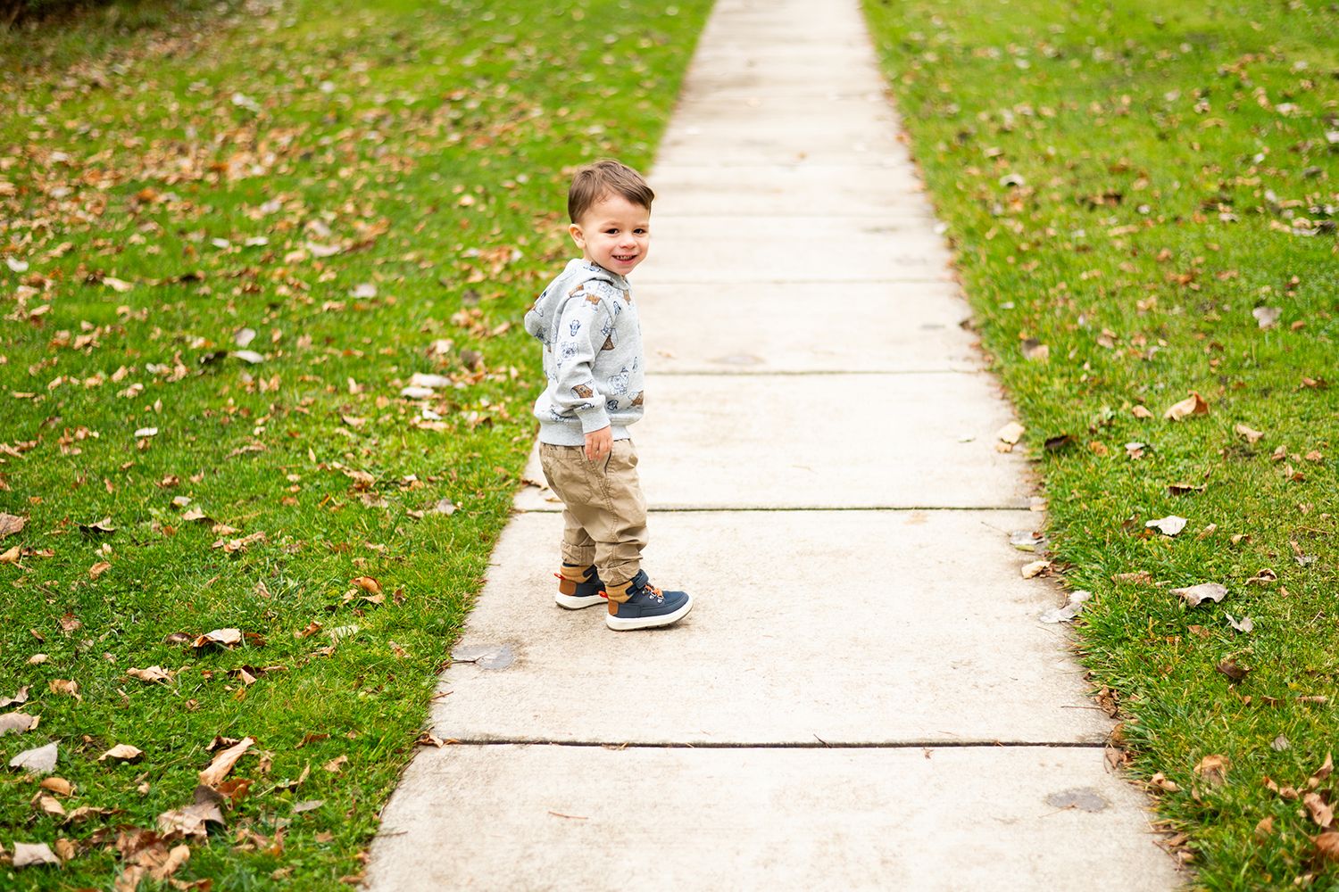 A small child walks along a concrete sidewalk with grass and fallen leaves on either side.