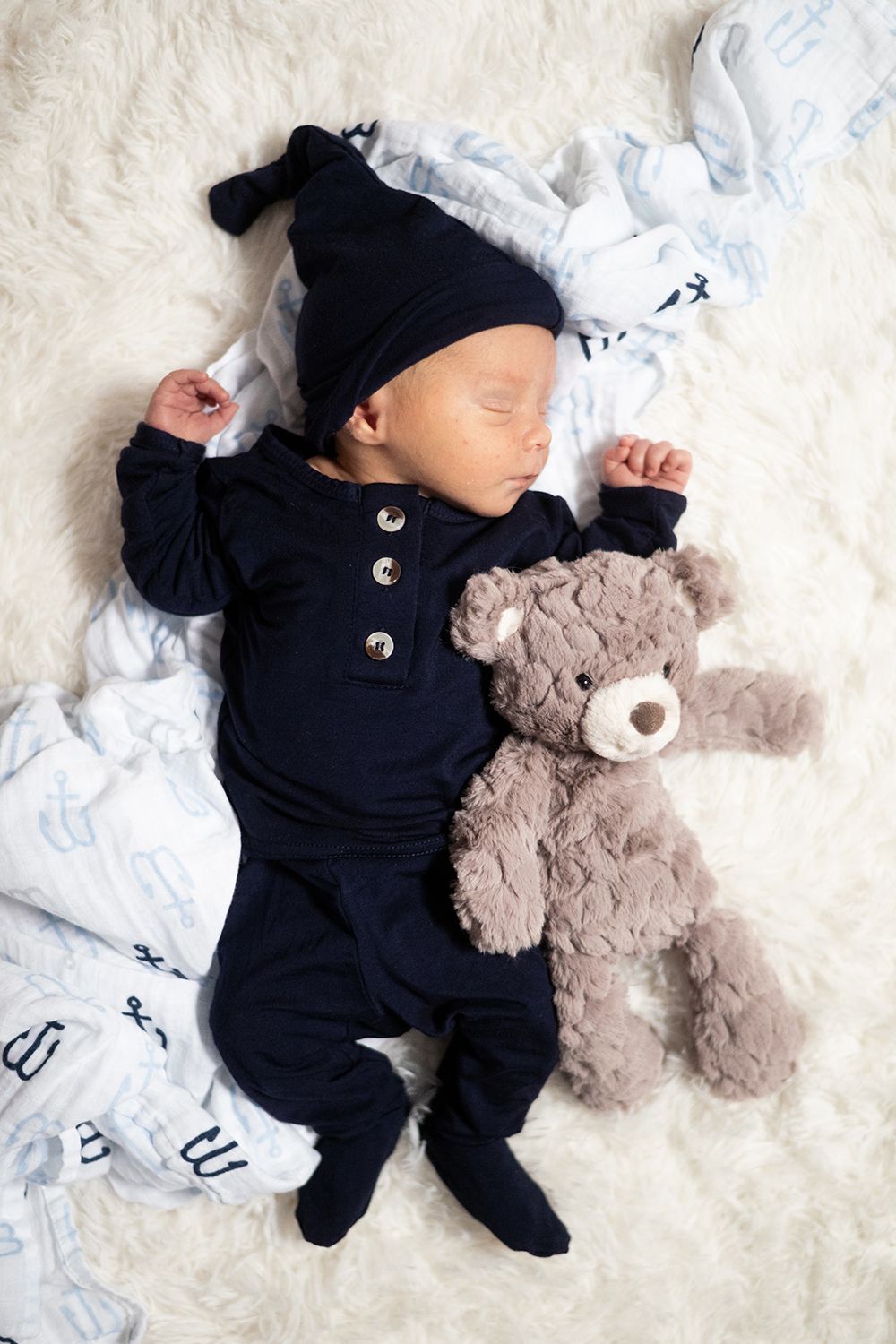 Sleeping baby wearing navy blue outfit and beanie lies on white blanket next to plush teddy bear.