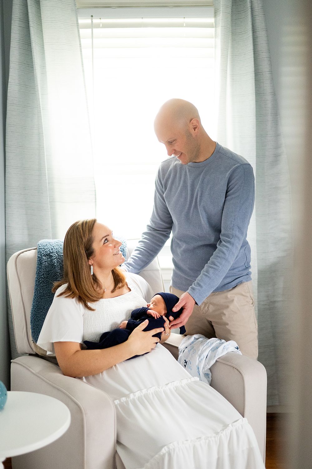 A loving couple shares an intimate moment in a bright living room with grey curtains while holding newborn baby.