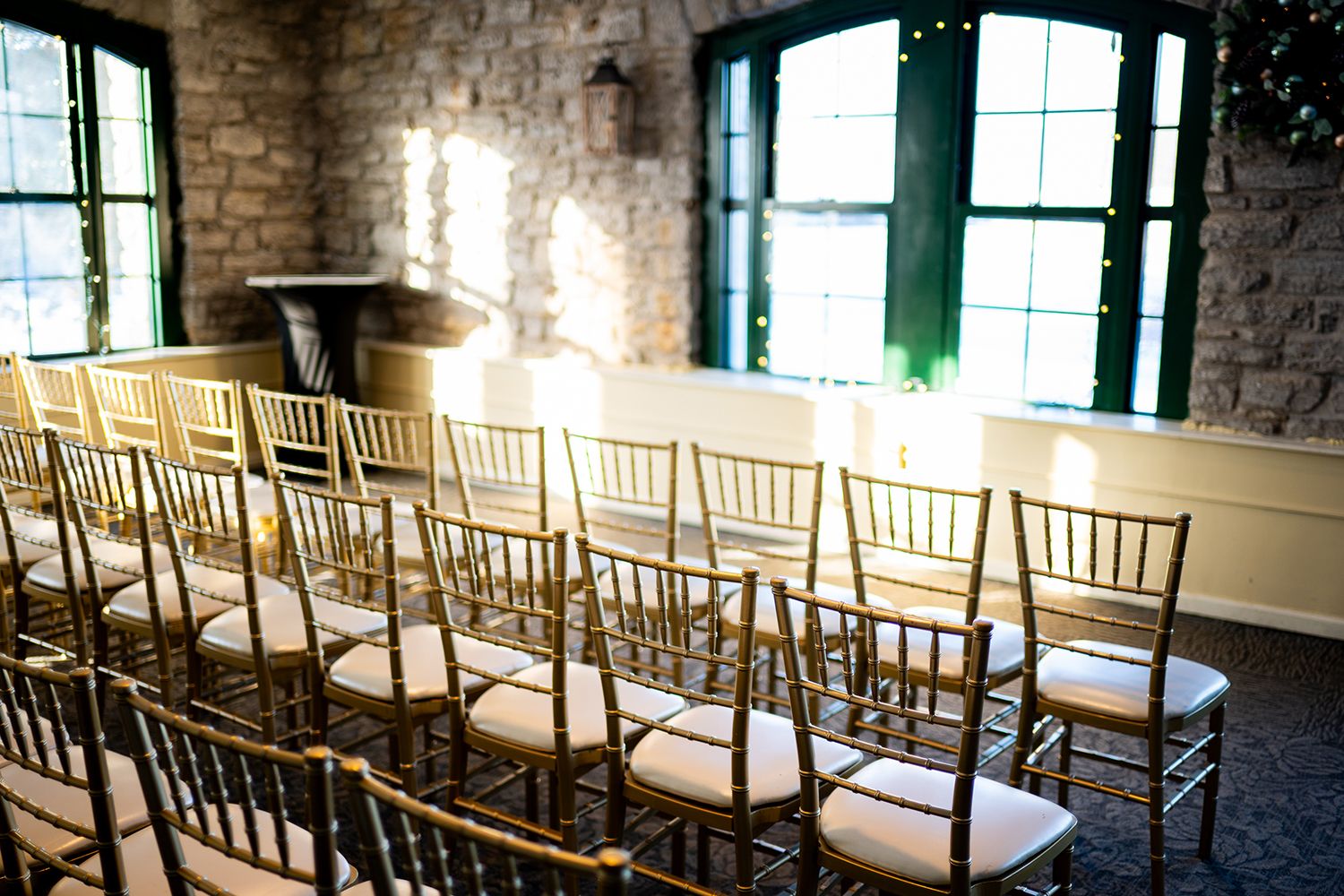 Rows of elegant gold chiavari chairs arranged for a ceremony in a rustic brick-walled room with sunlit windows.