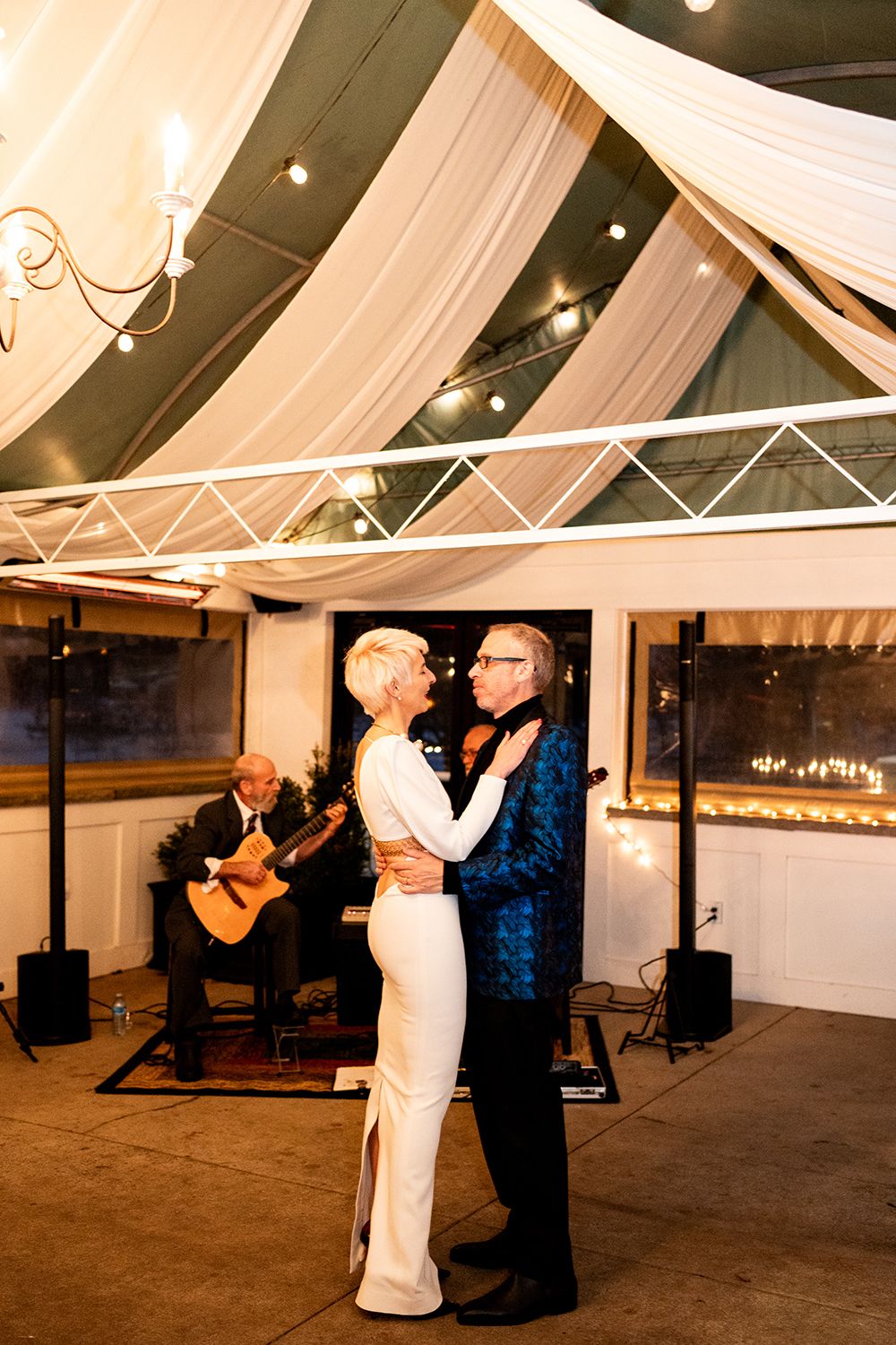 A couple dances in an elegant venue with draped fabric decor while musicians perform in the background.