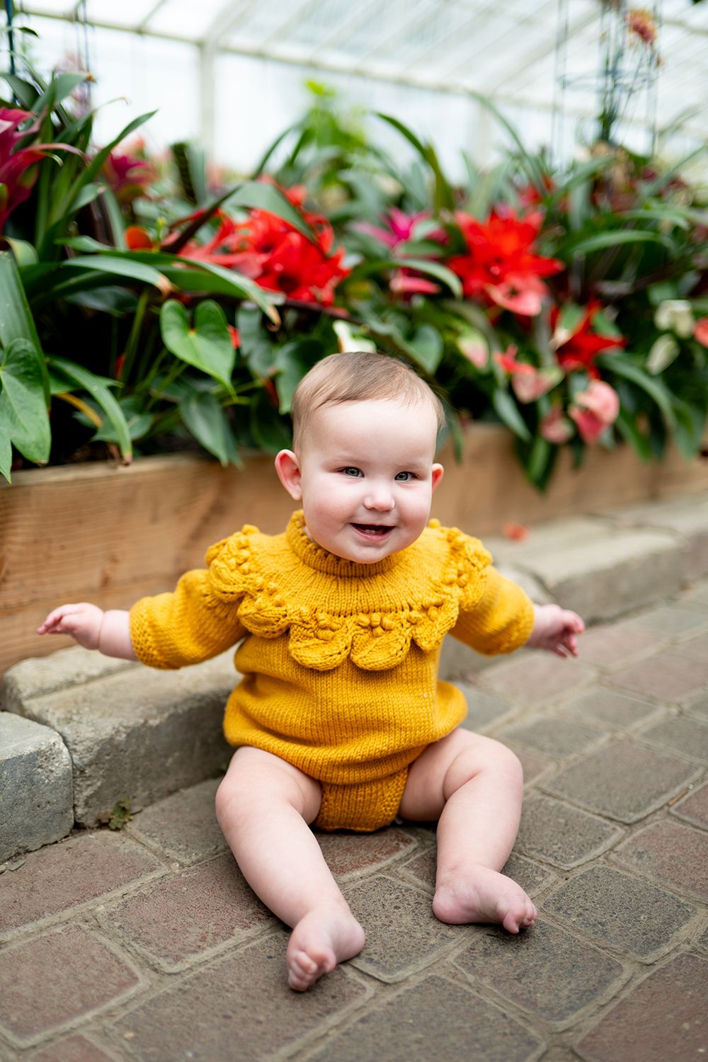 A small child wearing a bright yellow knit sweater sits on brick pavers in front of flowering plants in a greenhouse setting.