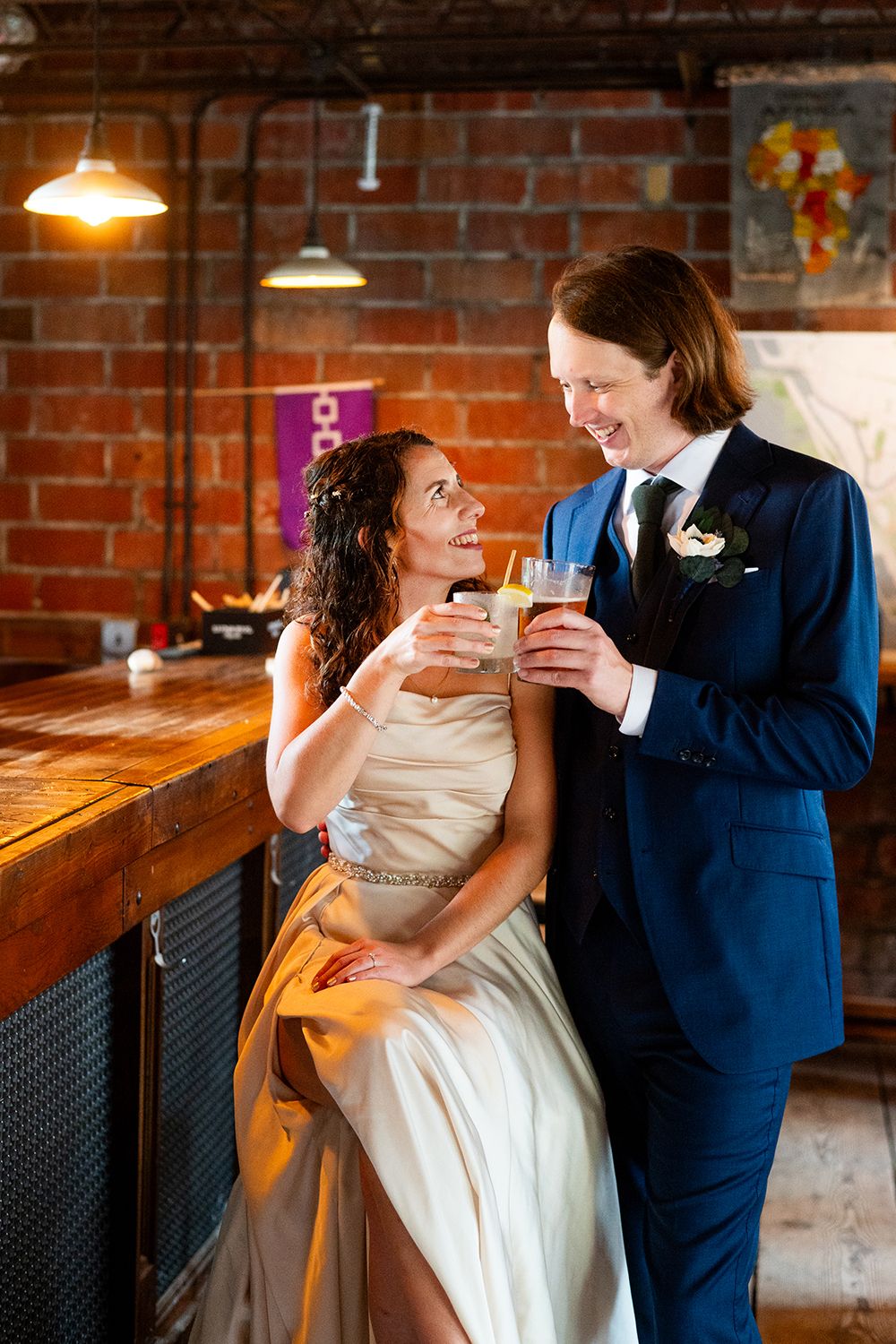 A couple in wedding attire share drinks at a rustic bar with exposed brick walls and pendant lighting.