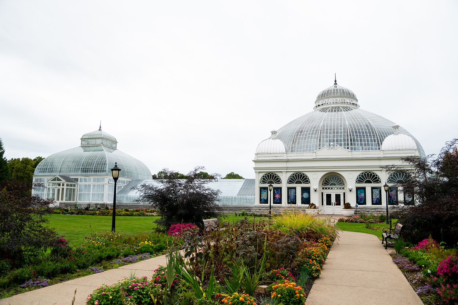 White conservatory building with glass domes surrounded by landscaped gardens on a cloudy day.