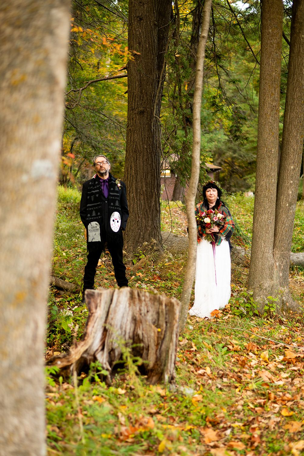 Two figures pose together in a wooded autumn setting with fallen leaves covering the ground.