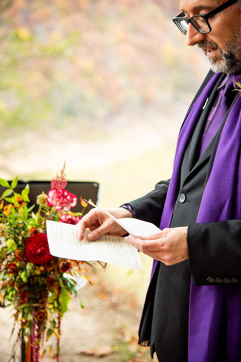 An officiant in black robes with a purple stole reads from a document during an outdoor ceremony.