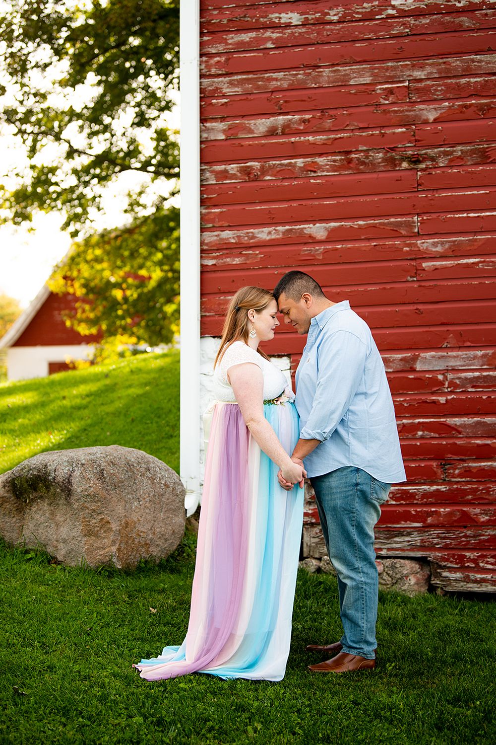 Romantic couple sharing tender moment against rustic red barn wall surrounded by lush green grass at sunset.