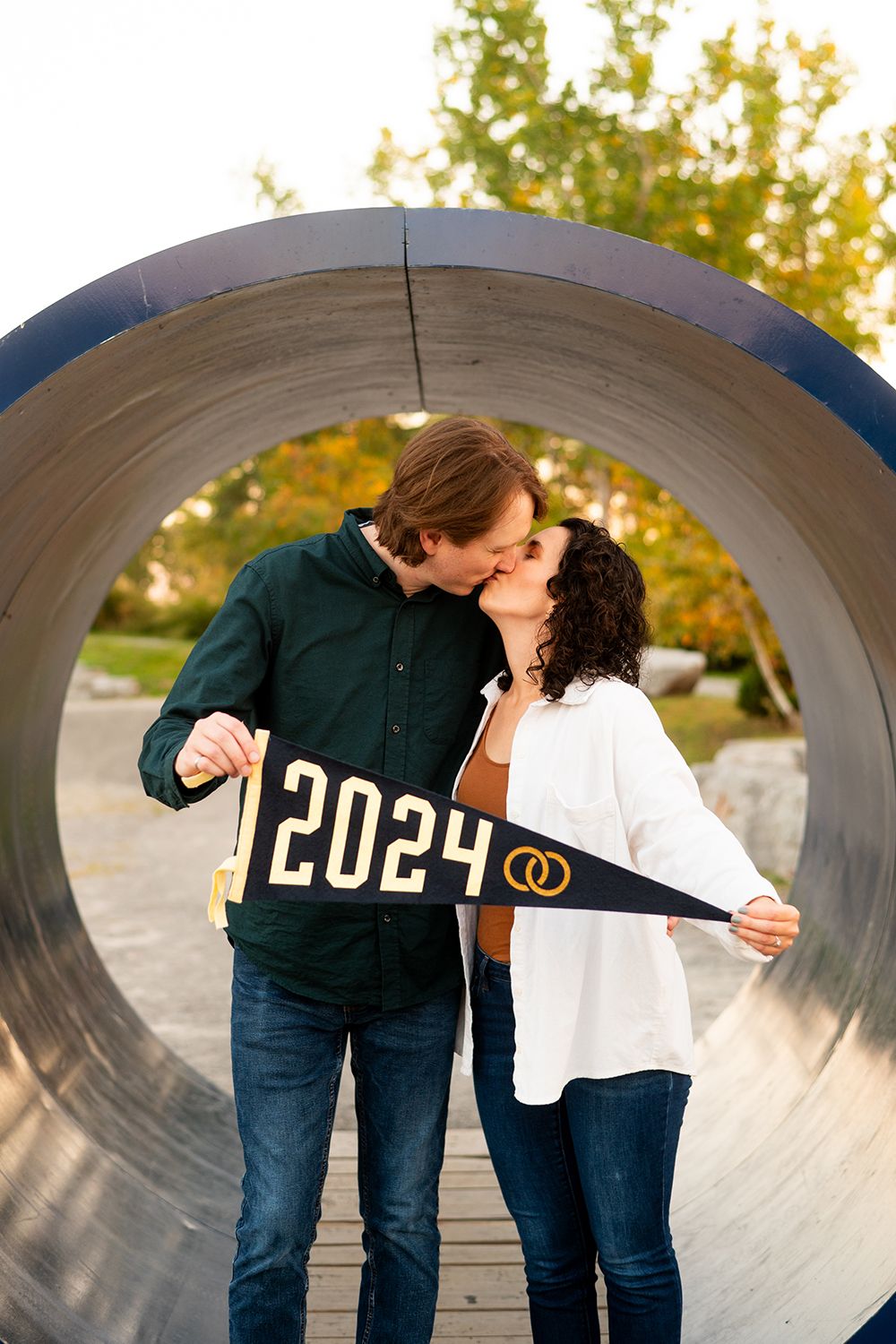 A couple sharing a romantic moment in a concrete tunnel while holding a 2024 pennant banner at sunset.