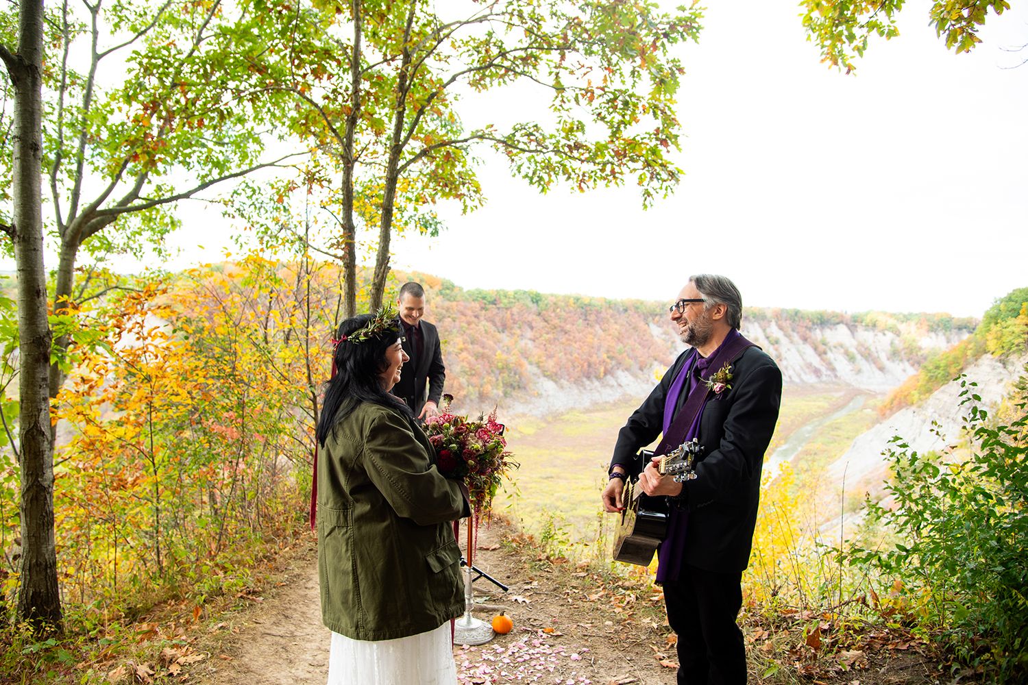 A couple stands on a cliff path during autumn with colorful fall foliage and a scenic overlook in the background.