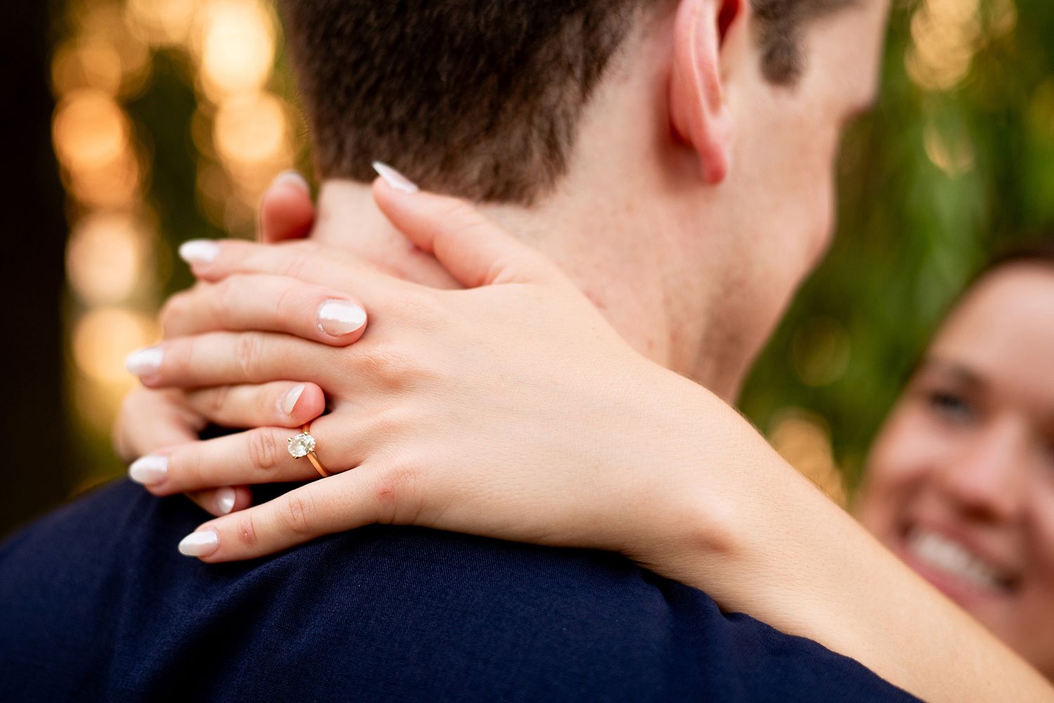 A close-up photo shows hands embracing around a neck with a sparkling engagement ring visible against a blurred background.