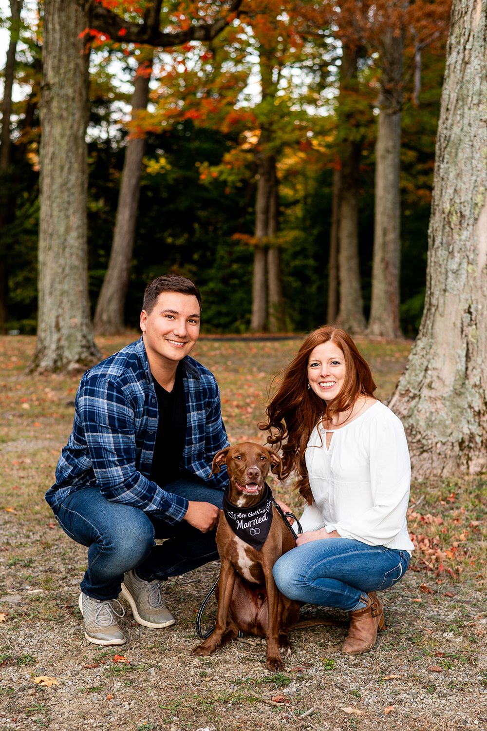 A couple poses with their brown dog in a wooded area during fall with trees in the background.