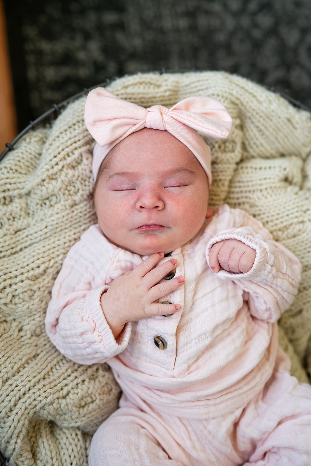 A sleeping newborn baby wearing pink clothing and a bow rests peacefully on a cream-colored knit blanket.