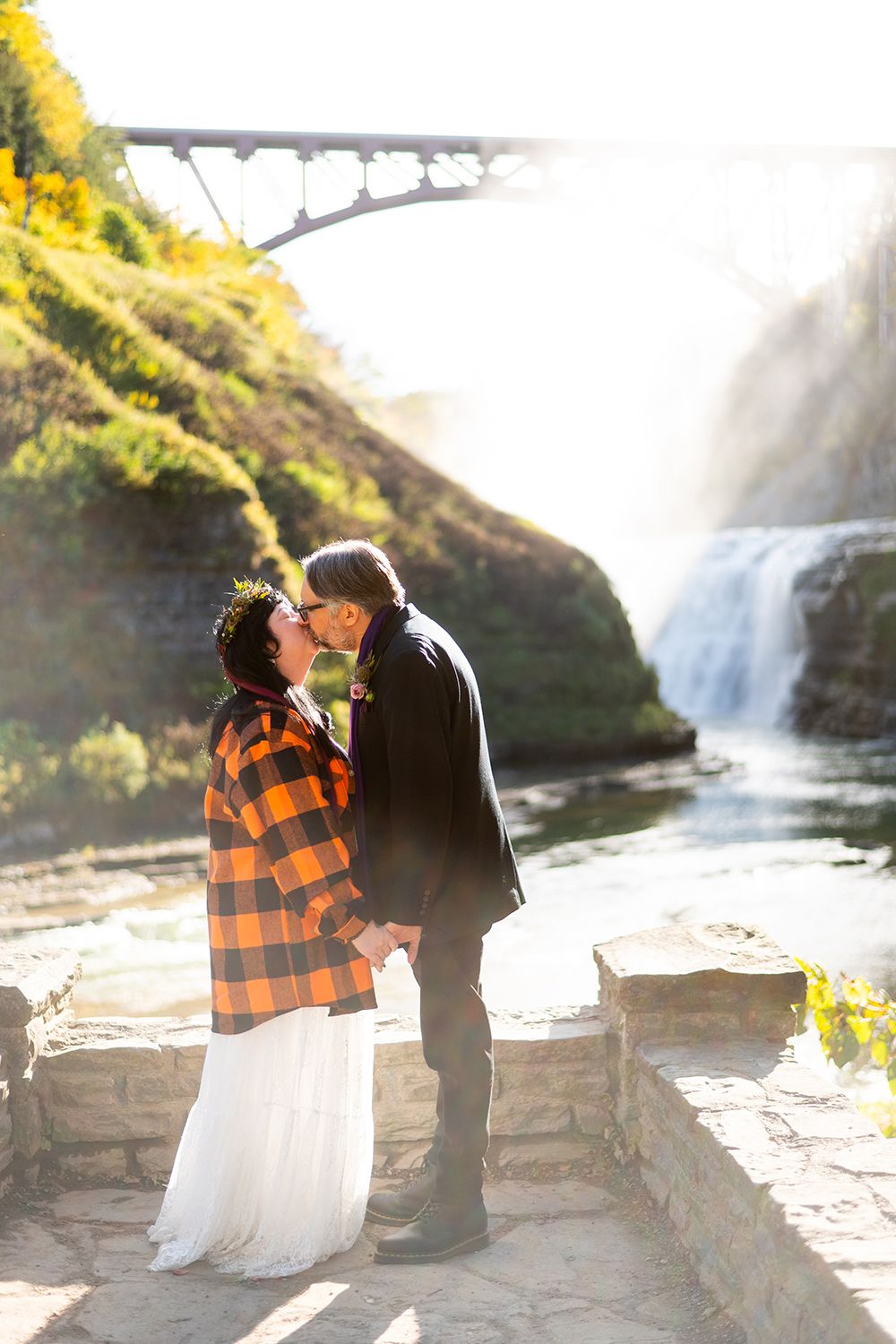 A couple in plaid and formal wear share romantic moments by a scenic waterfall during an engagement photoshoot.