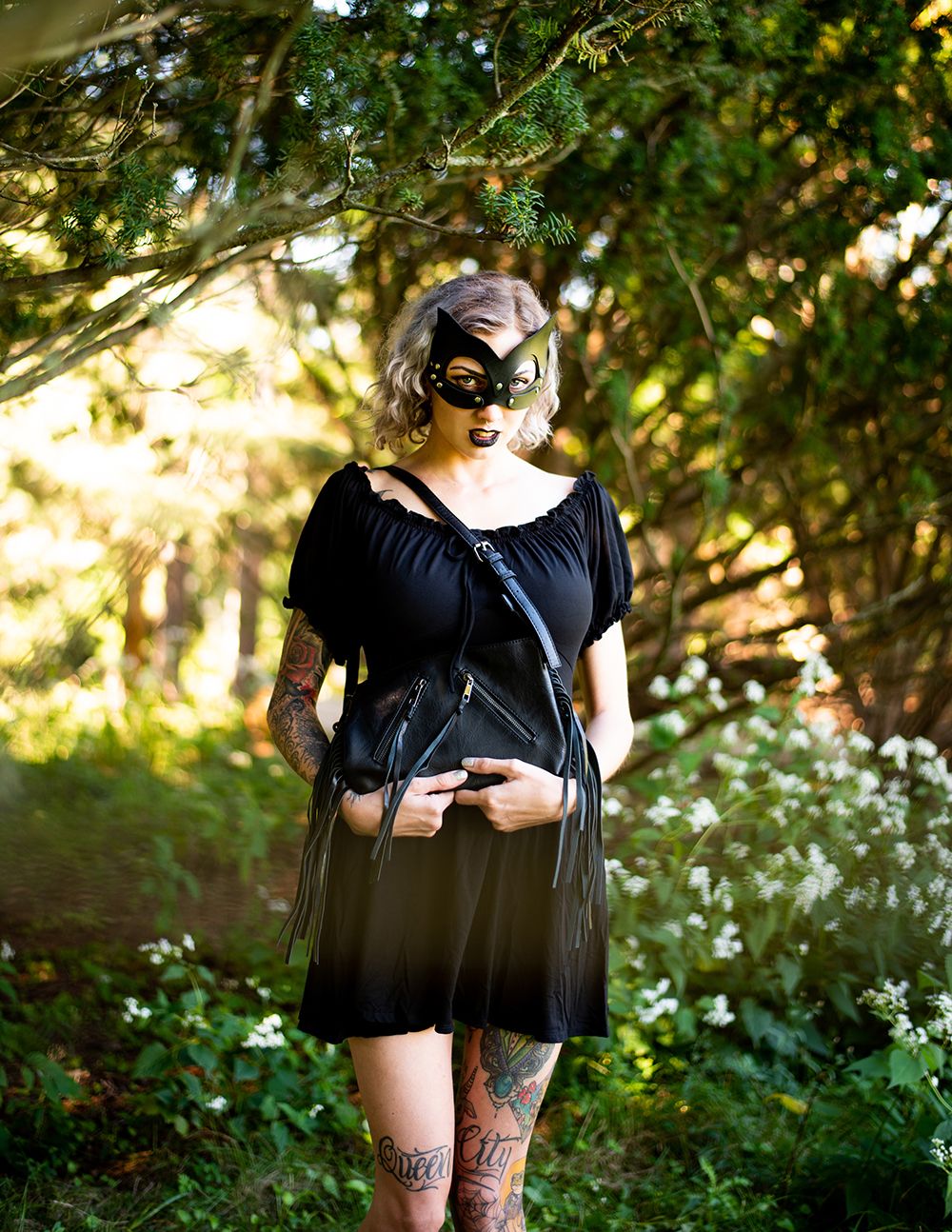 A figure in a black dress and cat-eye mask poses outdoors among wildflowers and greenery in golden sunlight.