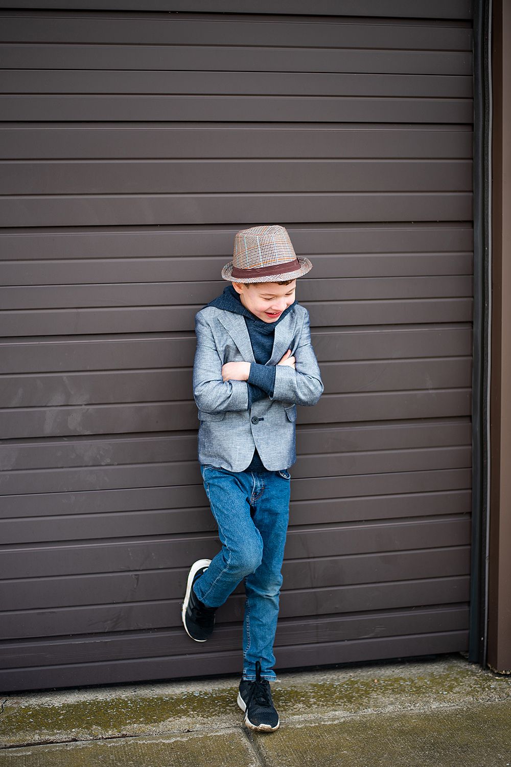 A young boy in a fedora hat and blue blazer leans casually against a gray garage door while striking a stylish pose.