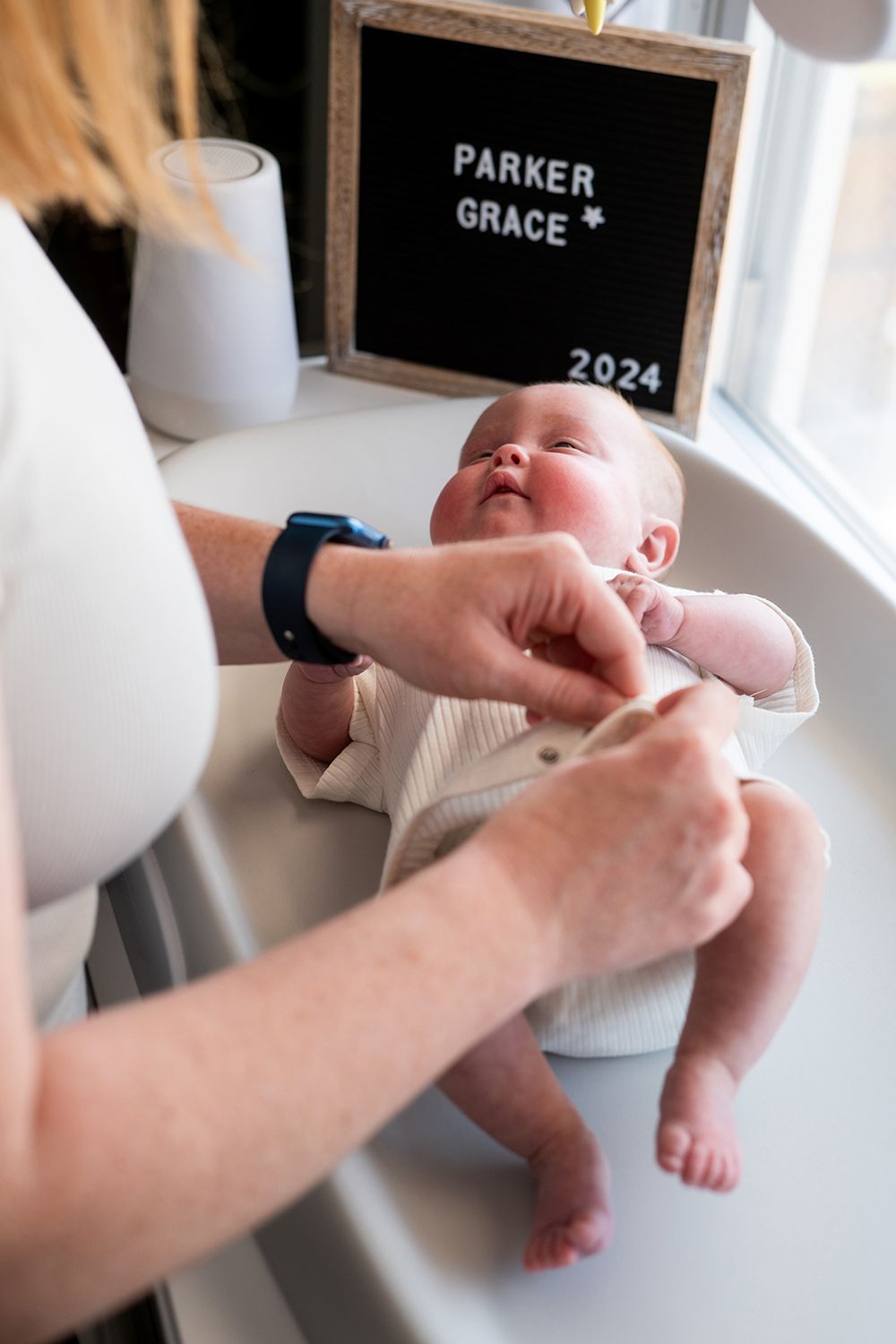 A baby in white clothing being changed on a white surface with a letterboard sign in the background.