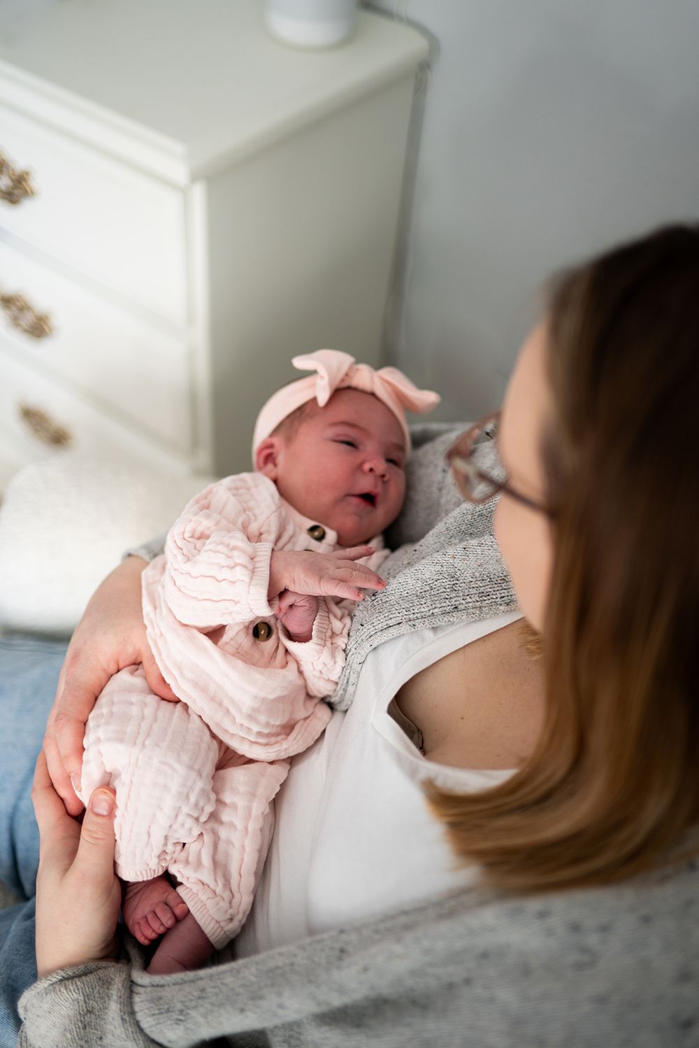 A baby wearing a pink bow and outfit lies against someone in a gray sweater.