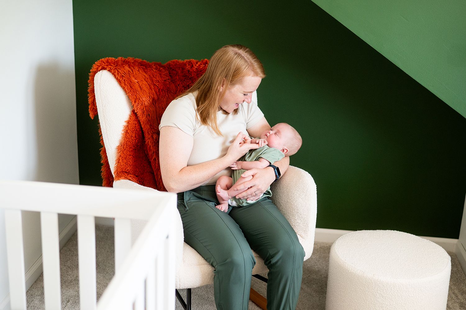 A parent sits in a cozy red armchair feeding a baby in a nursery with green walls.