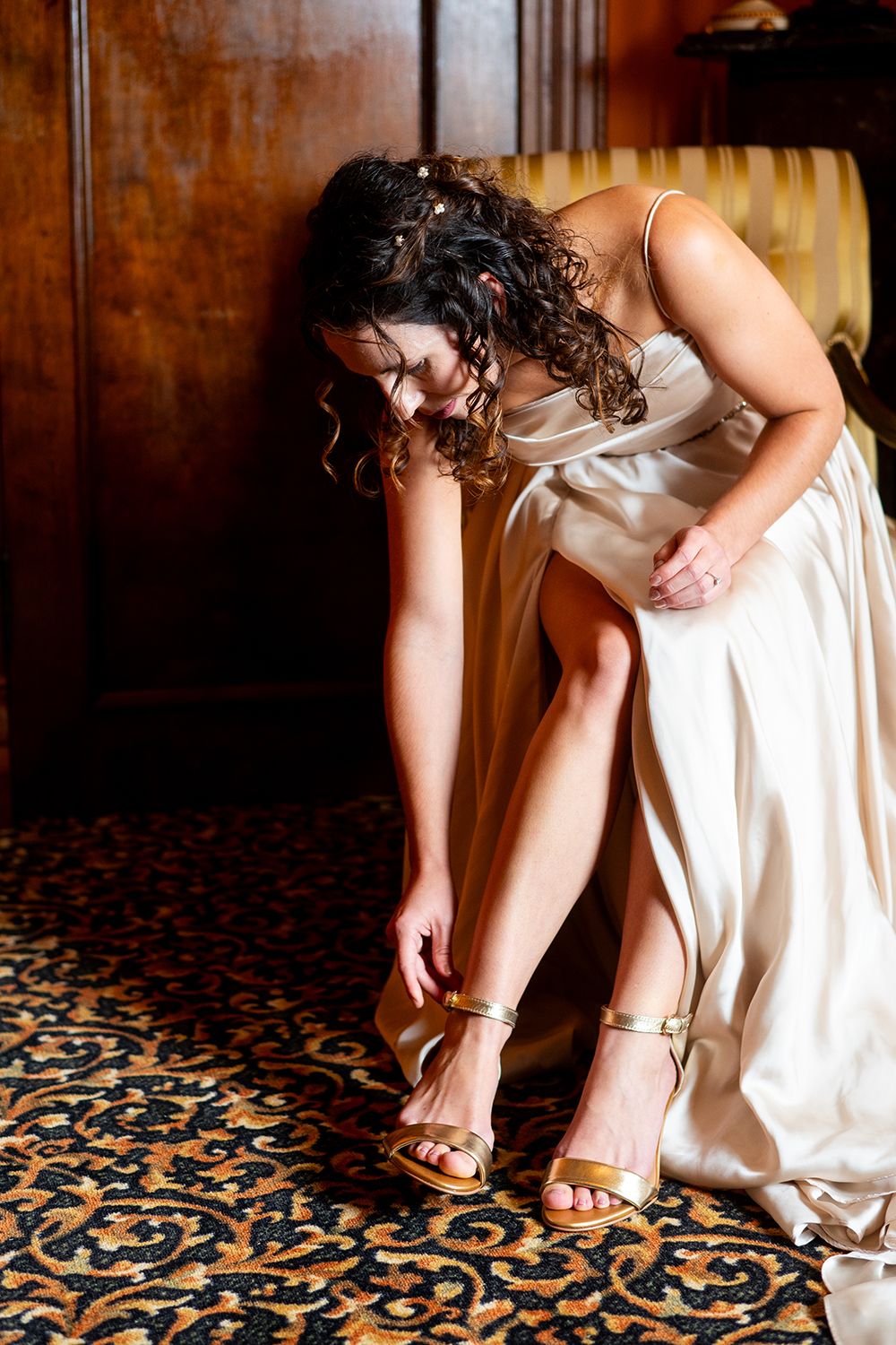 A woman in an ivory dress fastens gold sandals while seated on an ornate patterned carpet.