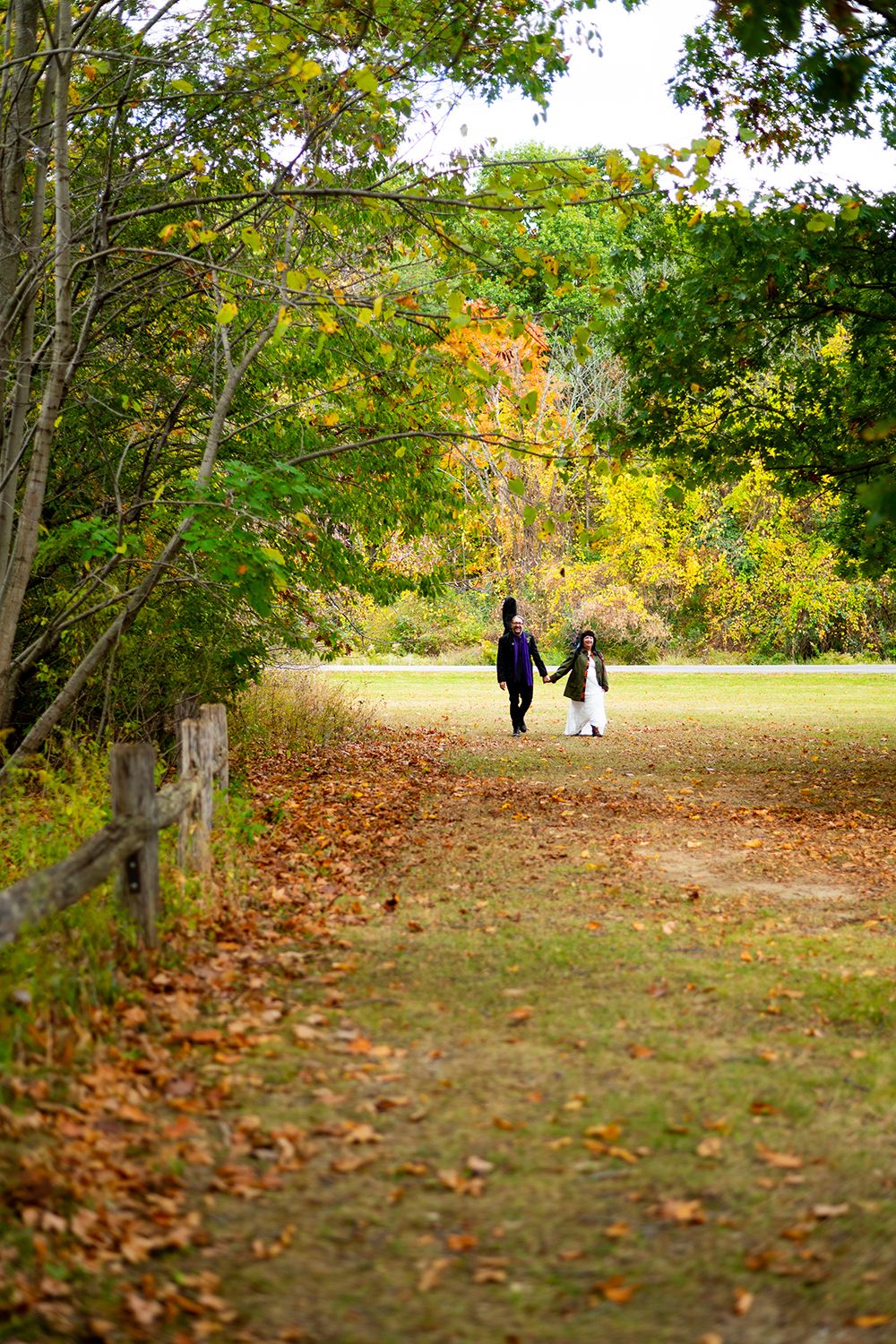 Two people walking hand in hand down a tree-lined autumn path with fallen leaves and golden foliage.